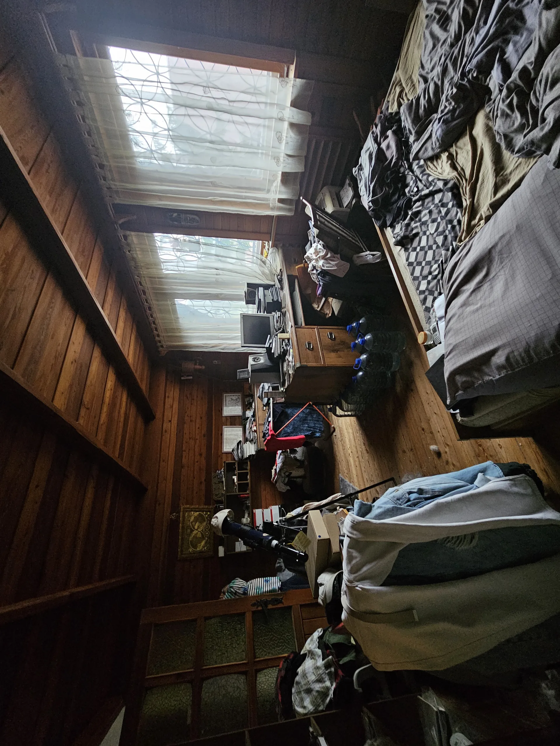 Wnętrze pokoju z drewnianymi ścianami i podłogą. Dwa okna z przezroczystymi zasłonami z geometrycznym wzorem. Niezrobione łóżko z różnymi kocami, w tym czarno-białym w kratę. Biurko z monitorem komputerowym, papierami i butelkami. Wiele ubrań, pudełek oraz innych przedmiotów rozłożonych na powierzchniach. Drewniana konstrukcja podłogi i ściany.