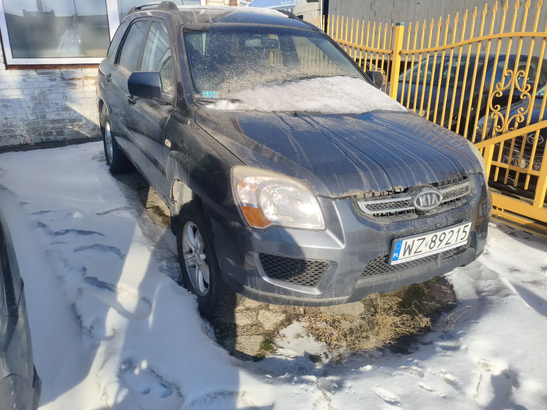 Samochód osobowy marki Kia, ciemnoszary, zaparkowany na śnieżnej powierzchni. Na masce i przedniej szybie widoczny śnieg. Tablica rejestracyjna z białym tłem i czarnymi literami WZ 89215 oraz niebieskim pasem z napisem PL po lewej stronie. W tle żółte metalowe ogrodzenie, fragment budynku z białą cegłą i oknem oraz inne zaparkowane pojazdy. Na ziemi ślady po stopach w śniegu.