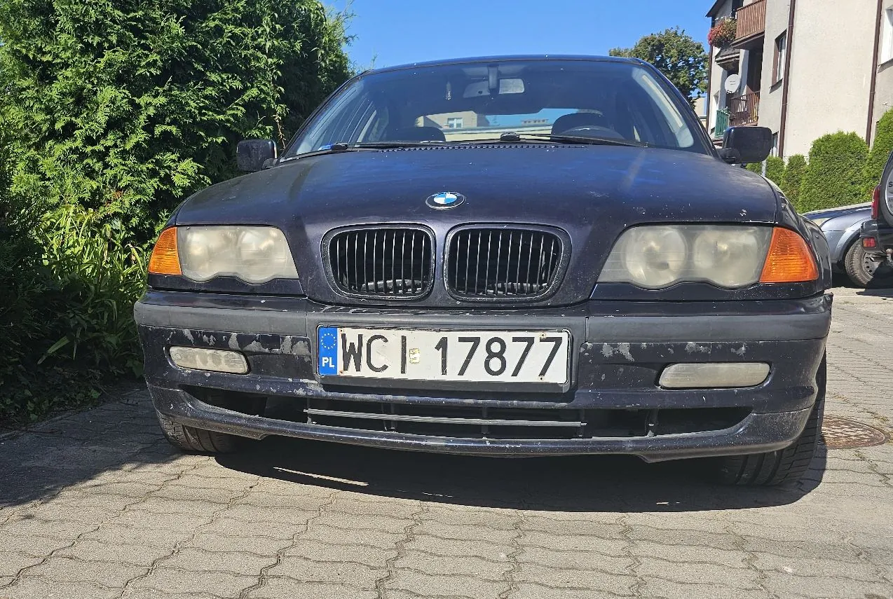 Widok z przodu na ciemnogranatowy samochód marki BMW zaparkowany na kostce brukowej. Pojazd posiada polską tablicę rejestracyjną o numerze WCI 17877. W tle widoczne są zielone krzewy oraz fragment budynku mieszkalnego pod bezchmurnym, niebieskim niebem.