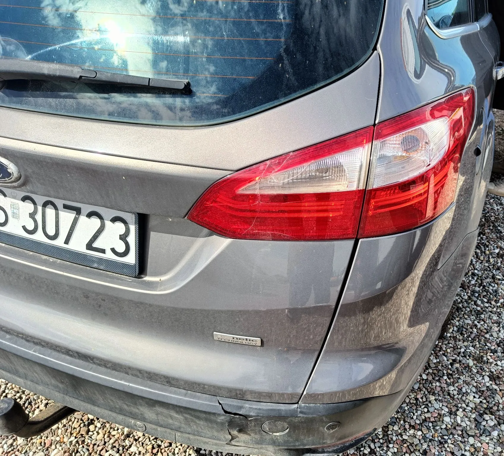 Zbliżenie na tył szarego samochodu. Widoczna jest prawa tylna lampa, fragment tablicy rejestracyjnej z numerem 30723 oraz napis 'netic' na klapie bagażnika. Na dolnym zderzaku widoczne jest pęknięcie i uszkodzenie lakieru. Samochód stoi na żwirowym podłożu.