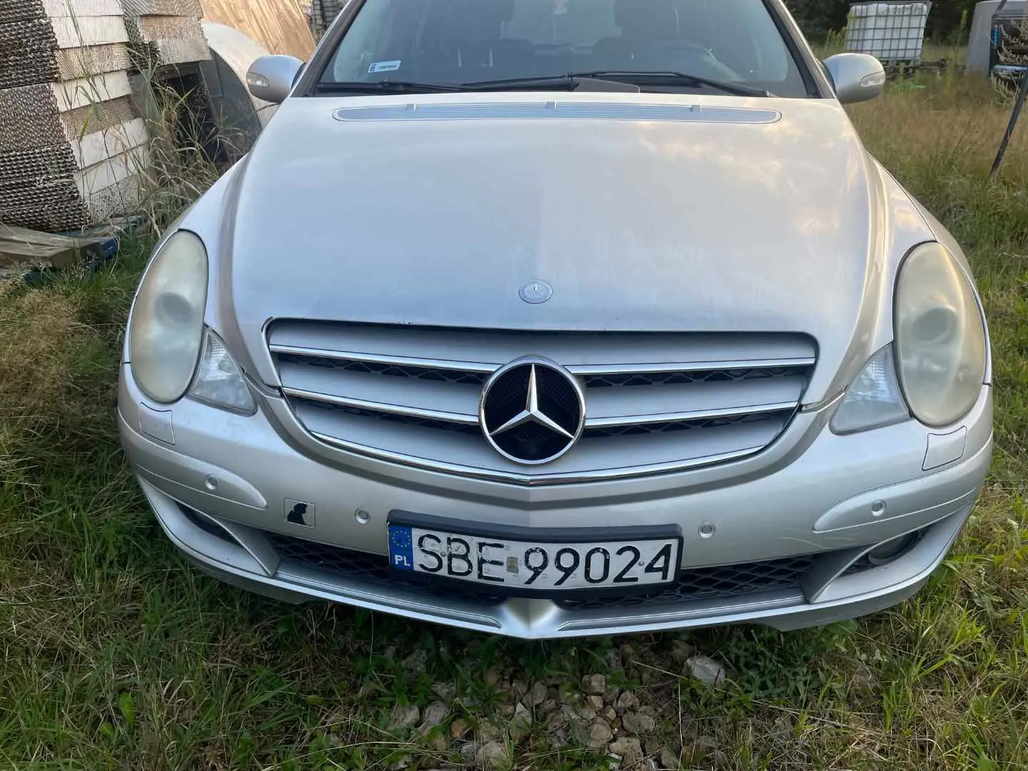 Srebrny samochód marki Mercedes-Benz widziany od przodu. Pojazd posiada charakterystyczne logo gwiazdy w centrum grilla oraz tablicę rejestracyjną o numerze SBE 99024. Samochód stoi na trawiastym podłożu, w tle widoczne są elementy ogrodzenia i roślinność.