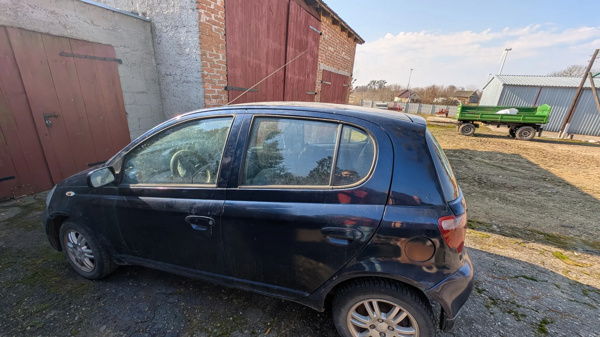 Czarny samochód osobowy typu hatchback zaparkowany na podwórku. W tle ceglany budynek z czerwonymi drewnianymi drzwiami garażowymi, zielony przyczepa oraz metalowy budynek. Podłoga pokryta żwirem i mchem, na niebie białe chmury.