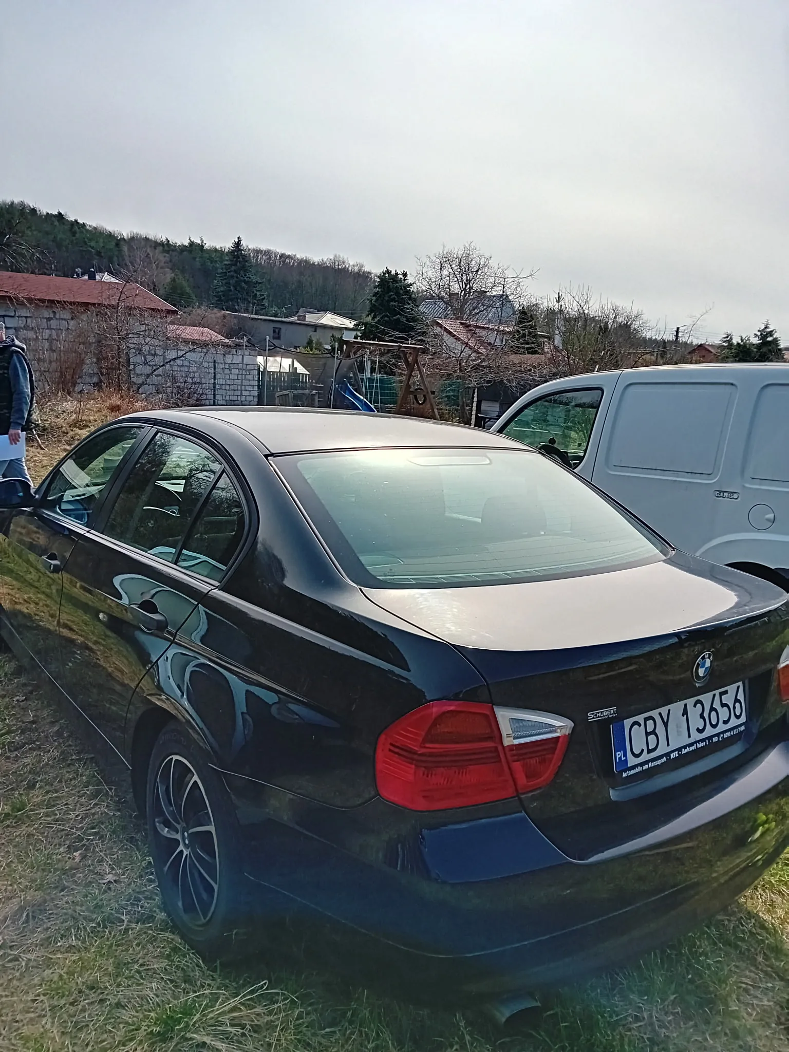 Czarny samochód osobowy marki BMW z tablicą rejestracyjną PL CBY 13656 zaparkowany na trawie. W tle biały furgon, domy z czerwonymi dachami, drzewa oraz osoba stojąca po lewej stronie. Niebo jest pochmurne, a na tle widoczne są wzgórza z lasem.