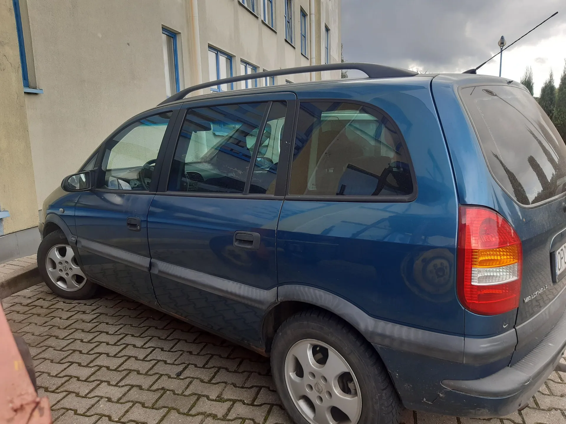 Niebieski samochód typu kombi zaparkowany na kostce brukowej obok jasnego budynku. Pojazd jest widoczny z profilu od strony pasażera, posiada relingi dachowe oraz szare listwy ochronne na drzwiach. W tle widać ścianę budynku z niebieskimi ramami okien oraz zachmurzone niebo.
