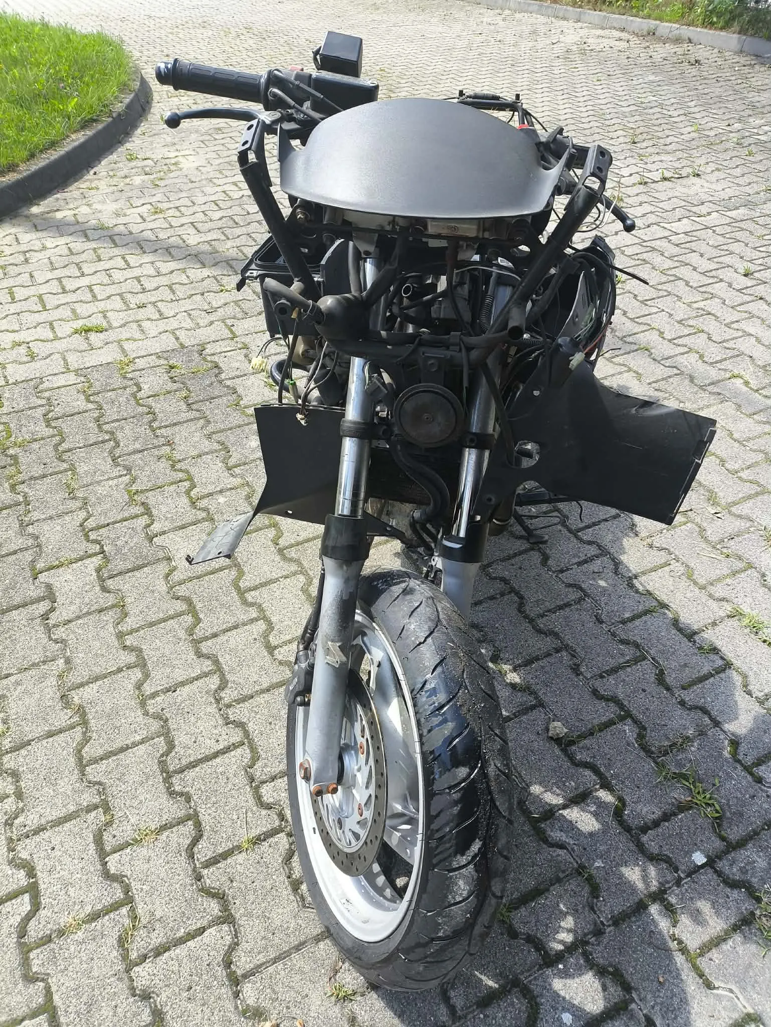 Zdjęcie przedstawiające przód rozmontowanego motocykla, ustawionego na kostce brukowej. Motocykl nie posiada owiewek ani reflektora, widoczna jest czarna rama, kierownica, przedni widelec oraz koło z oponą. Nad widelcem znajduje się szara, matowa osłona wykonana z tworzywa.