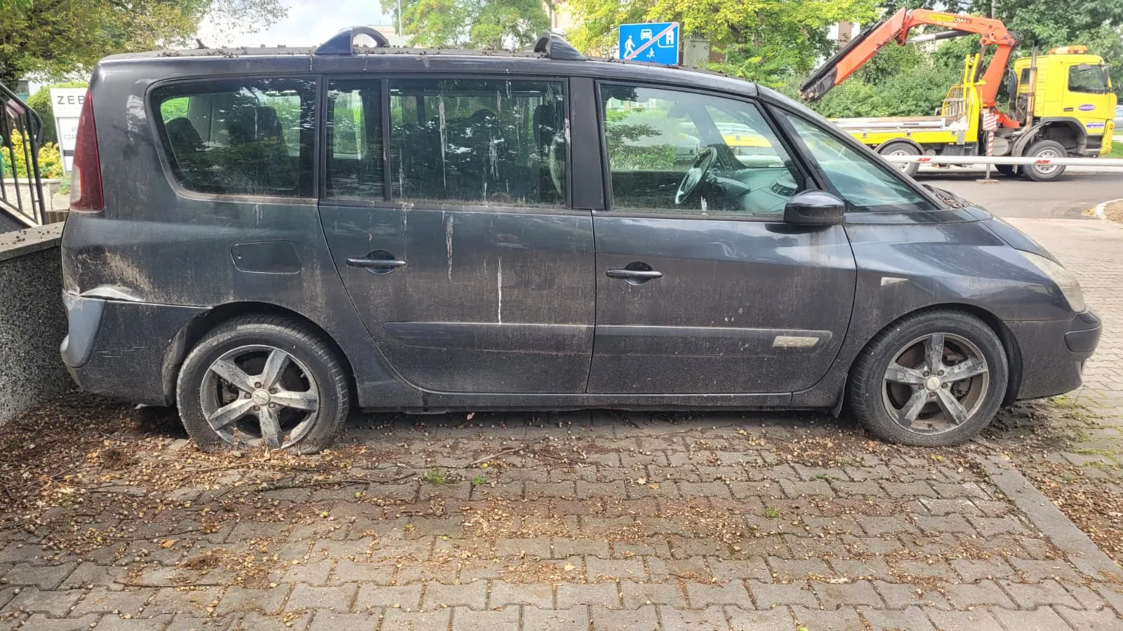 Profil brudnego, ciemnoszarego samochodu typu minivan zaparkowanego na kostce brukowej. Pojazd ma zamontowane relingi dachowe. W tle widoczny jest żółty samochód ciężarowy z dźwigiem oraz zielone drzewa.