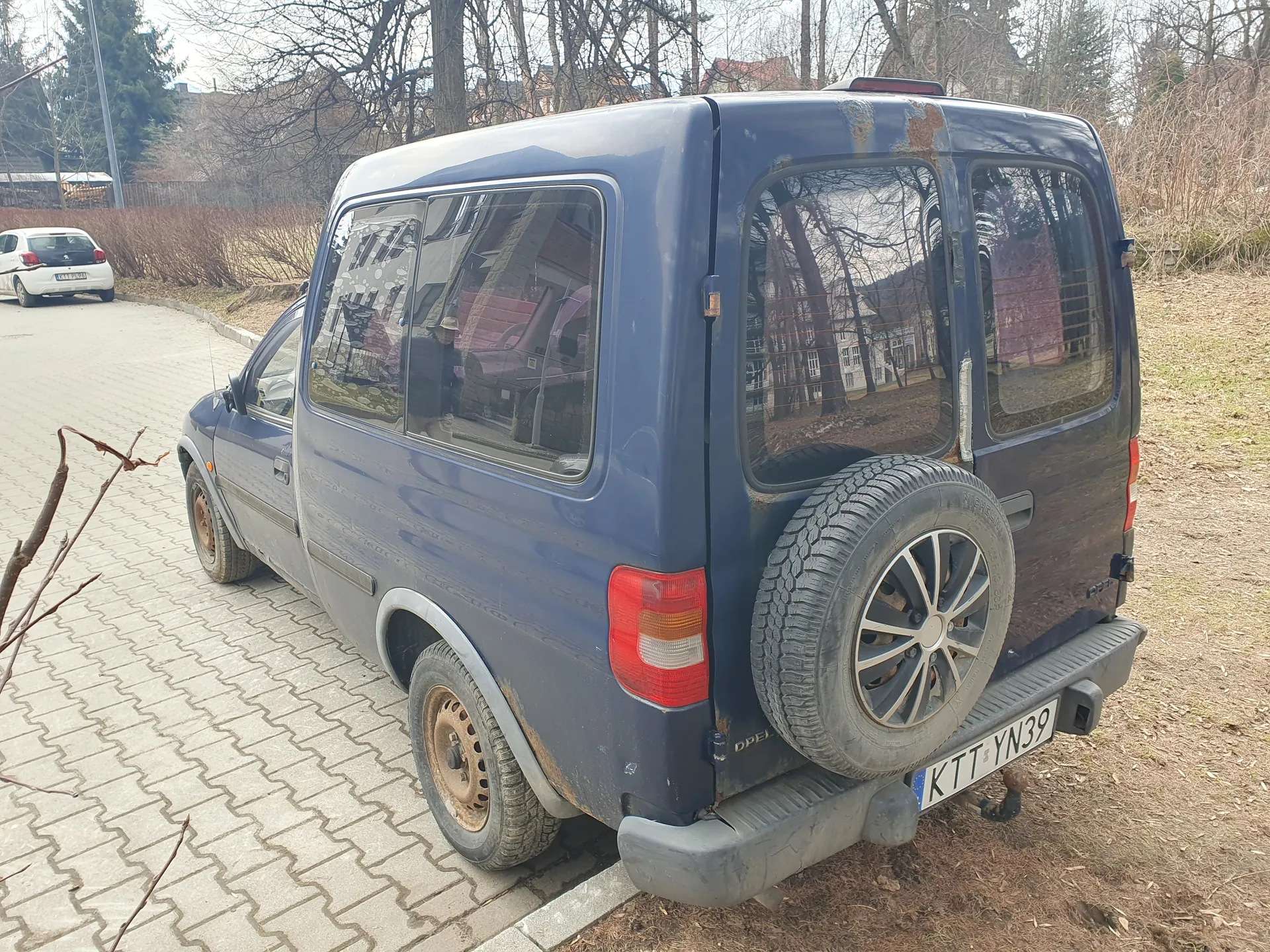 Niebieski furgon marki Opel z zapasowym kołem zamontowanym na tylnej klapie. Widoczny jest polski numer rejestracyjny KTT YN39. Furgon ma ślady rdzy na nadwoziu i brudne felgi. Pojazd stoi na brukowanej powierzchni, w tle biały samochód, suche krzewy, drzewa bez liści oraz budynki. Na tle widoczne są również suche trawy i fragment drogi.