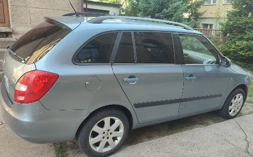 Srebrny samochód typu kombi zaparkowany na betonowym podjeździe. Widok z boku i z tyłu pojazdu. Samochód posiada relingi dachowe, przyciemniane tylne szyby oraz czarne listwy ochronne na drzwiach.