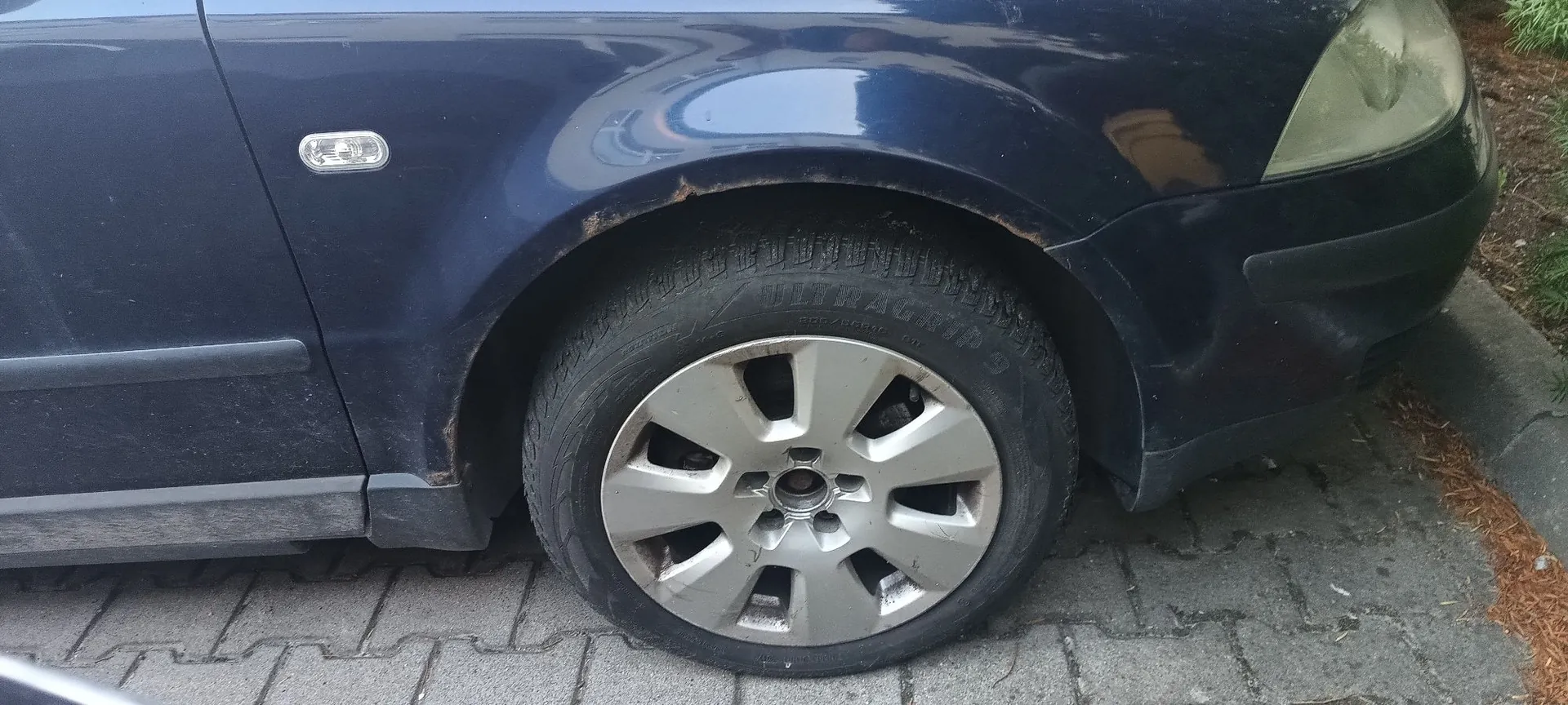 Zbliżenie na przednie prawe koło ciemnoniebieskiego samochodu zaparkowanego na kostce brukowej. Widoczna jest felga aluminiowa z pięcioma szerokimi ramionami oraz opona marki Vitragrip. Nad nadkolem widoczne są ślady korozji i rdzy na lakierze.