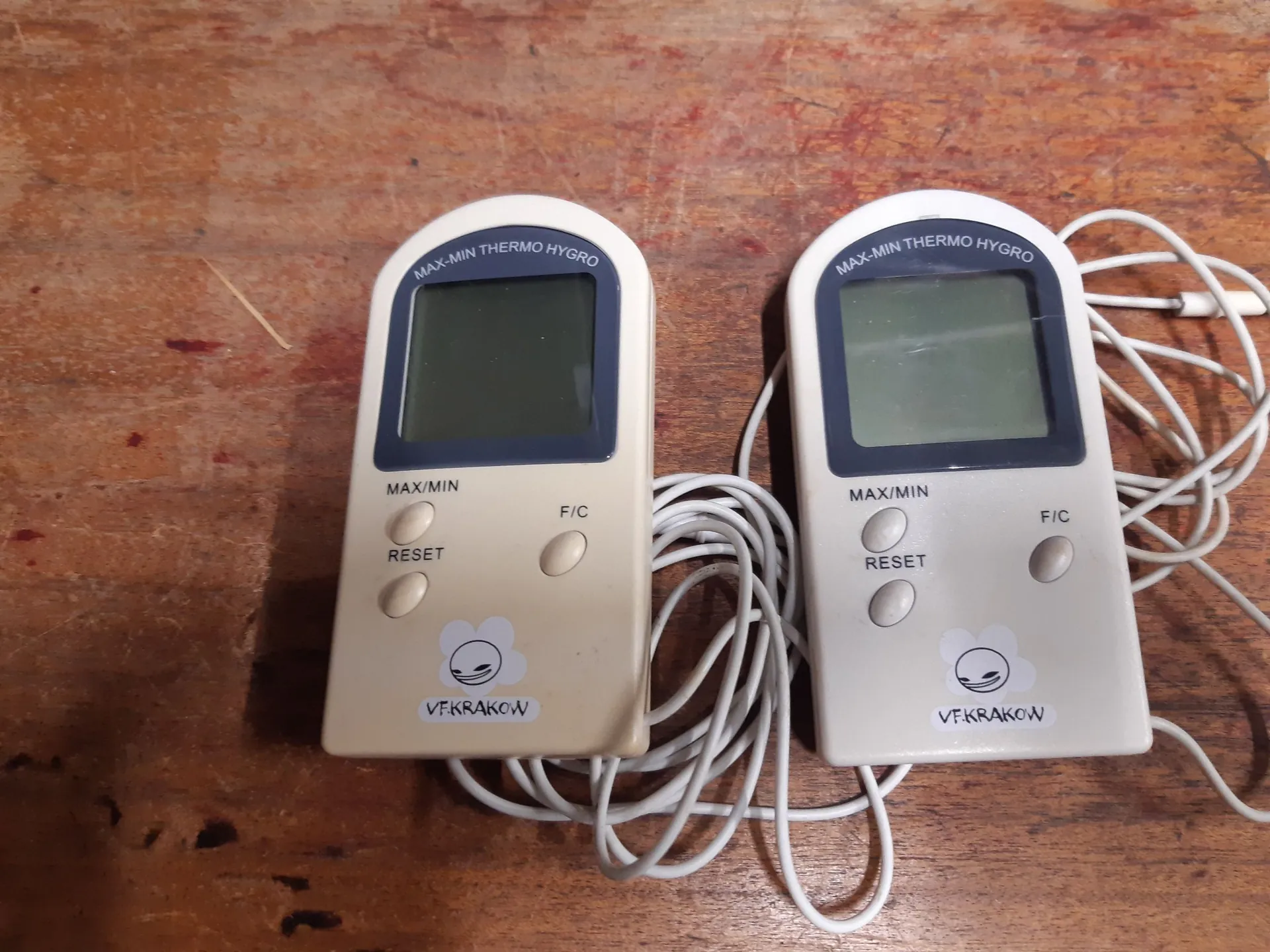 Dwa identyczne, białe cyfrowe termohigrometry z wyświetlaczami LCD i zewnętrznymi czujnikami na białych przewodach, leżące obok siebie na drewnianym blacie. Na górze każdego urządzenia widnieje napis 'MAX-MIN THERMO HYGRO'. Poniżej ekranu znajdują się trzy przyciski: 'MAX/MIN', 'RESET' oraz 'F/C'. Na dole obudowy znajduje się logo z chmurką i napisem 'VF.KRAKOW'.
