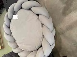 Okragly poduszka w kolorze jasnoszarym z plecionym brzegiem. Na poduszce widoczny bialy etykietka. Tlo stanowi beżowa tkanina, prawdopodobnie mebel lub pled, oraz czesc ciemnego przedmiotu w lewym dolnym rogu.