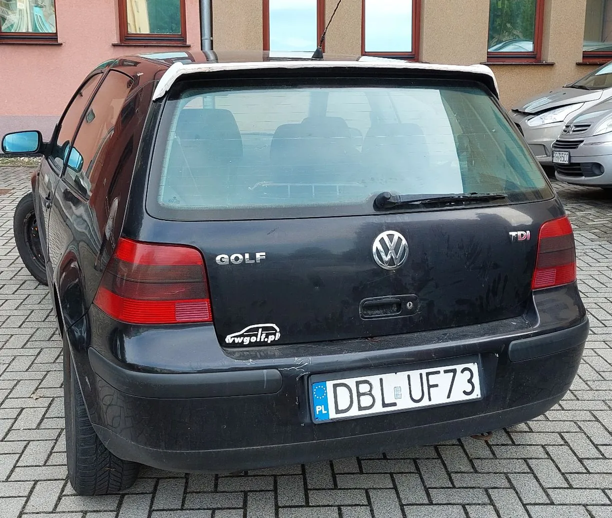 Tylny widok czarnego samochodu marki Volkswagen Golf w wersji kombi. Na klapie bagażnika widoczne są napisy 'GOLF' oraz 'TDI' oraz logo Volkswagena. Samochód posiada polską tablicę rejestracyjną o numerze 'DBL UF73'. Poniżej napisu Golf znajduje się naklejka z adresem 'vwgolf.pl'. Auto zaparkowane jest na kostce brukowej przed budynkiem o beżowej elewacji.