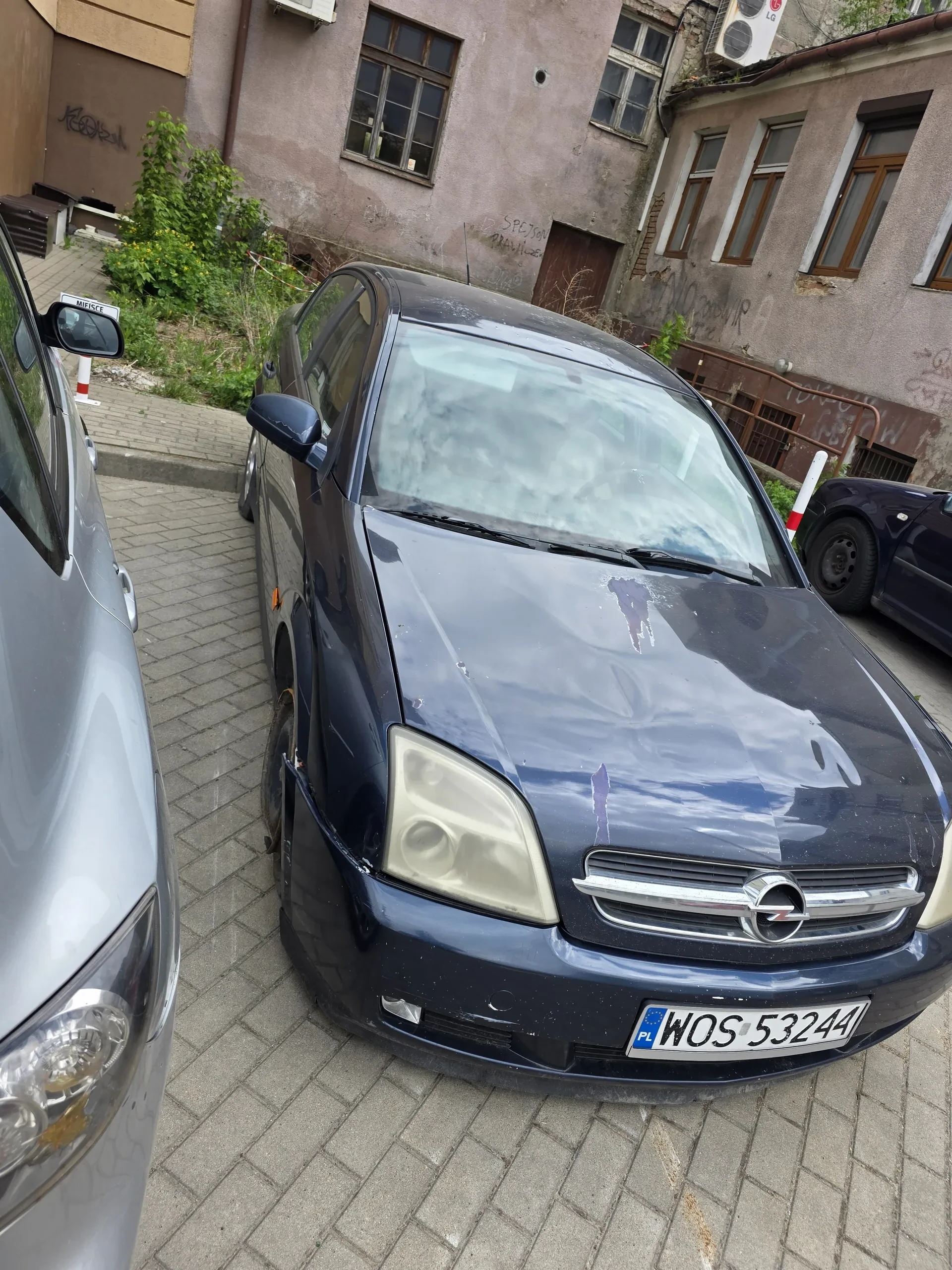 Ciemnoniebieski samochód marki Opel zaparkowany na brukowanej powierzchni. Widoczna tablica rejestracyjna z pionowo umieszczonymi literami i cyframi WOS 5324. W tle budynki z oknami, graffiti na ścianach, klimatyzator oraz inne zaparkowane samochody. Na pierwszym planie częściowo widoczny srebrny samochód.