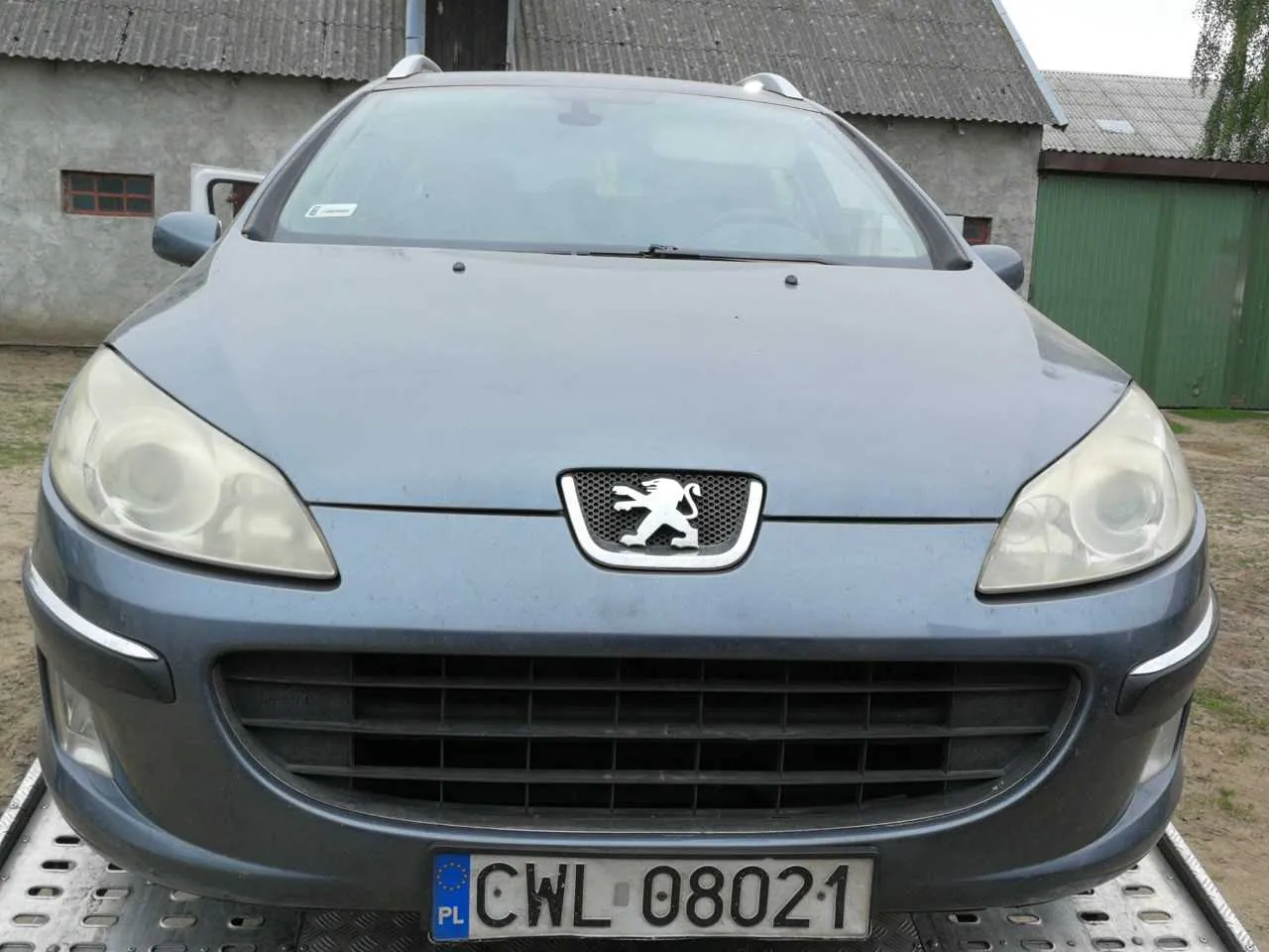 Przód szarego samochodu marki Peugeot z widocznym logo lwa na atrapie chłodnicy. Tablica rejestracyjna z napisem 'PL CWL 08021' umieszczona na dole. Samochód stoi na metalowej platformie, na tle budynku z blachą falowaną i zielonej bramy. Widoczne są reflektory, przednia szyba oraz fragmenty otoczenia z ziemią i trawą.