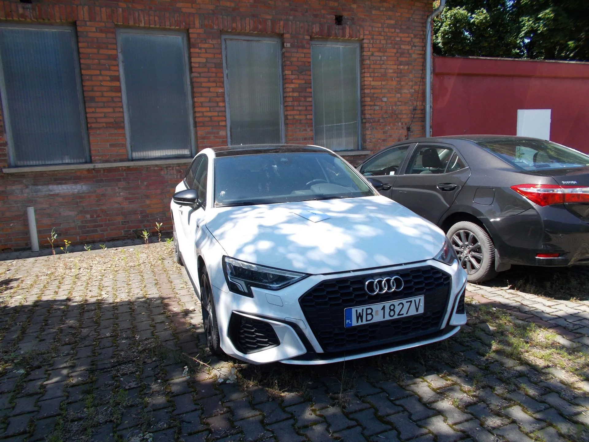 Biały samochód marki Audi zaparkowany na kostce brukowej przed budynkiem z czerwonej cegły z czterema wysokimi, wąskimi oknami. Obok stoi czarny samochód. Na przednim zderzaku białego auta widoczna jest tablica rejestracyjna o treści WB 1827V.