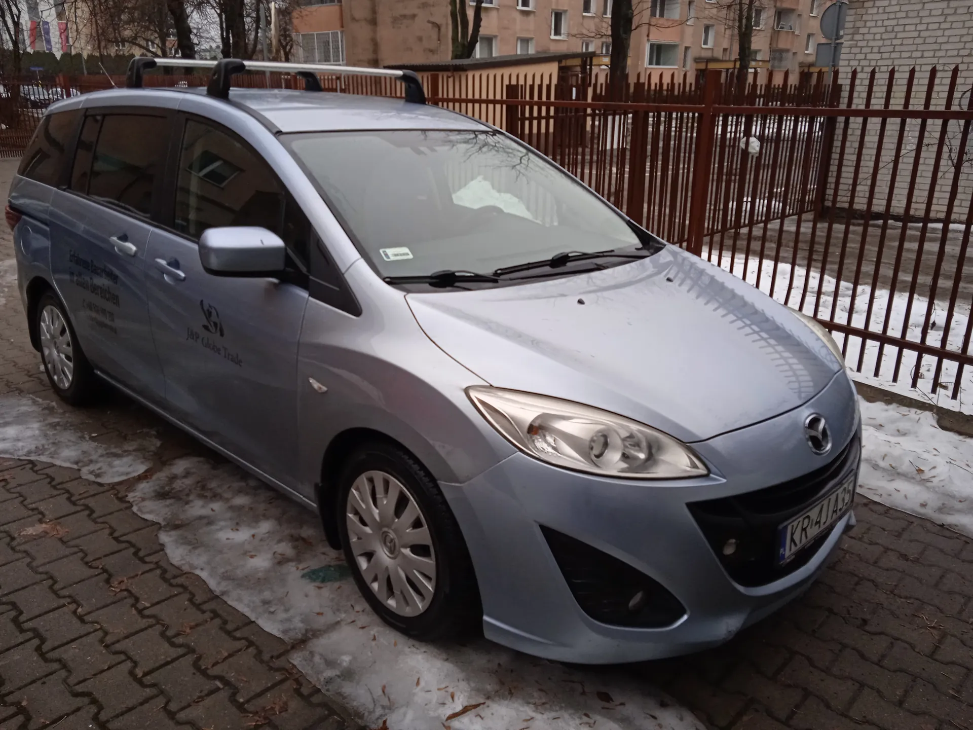 Srebrny samochód osobowy marki Mazda z dachowym bagażnikiem, zaparkowany na brukowanej powierzchni z resztkami śniegu. Na boku pojazdu widoczny napis 'J&P Globe Trade' oraz niemiecki tekst 'Erfahrene Berater in allen Bereichen'. Tablica rejestracyjna 'KR-4JA35'. W tle ceglany budynek, metalowe ogrodzenie i drzewa bez liści.