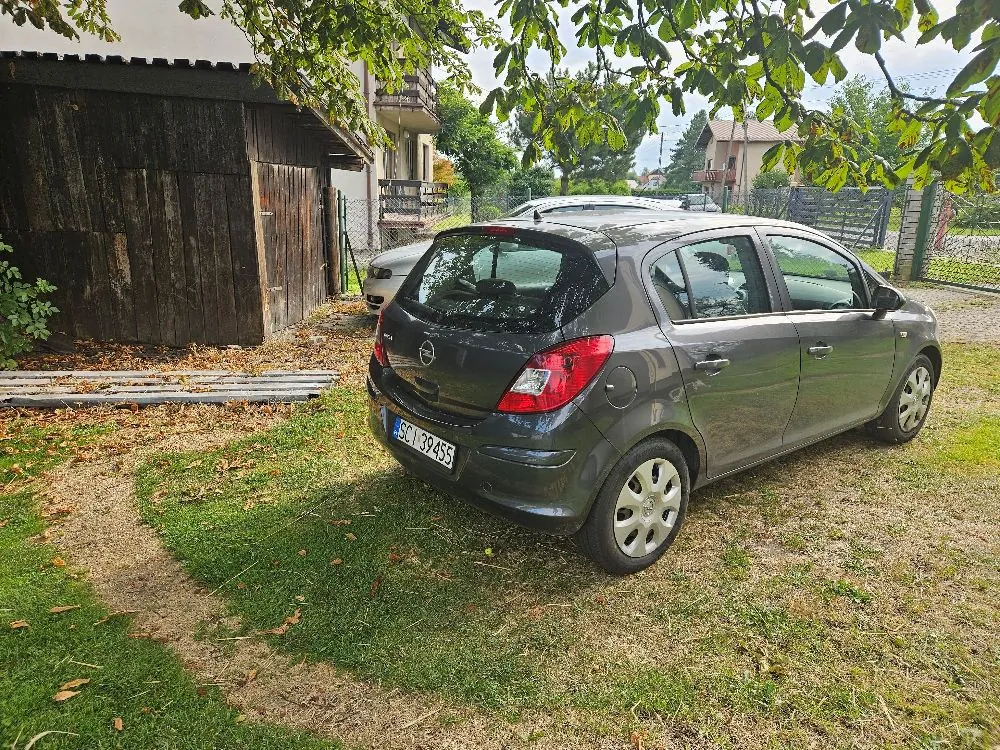 Szary samochód marki Opel Corsa zaparkowany na trawiastym podjeździe obok drewnianego budynku. Samochód ma tablicę rejestracyjną o numerze SCI 39455. W tle widoczne są drzewa, krzewy oraz fragmenty innych budynków mieszkalnych.
