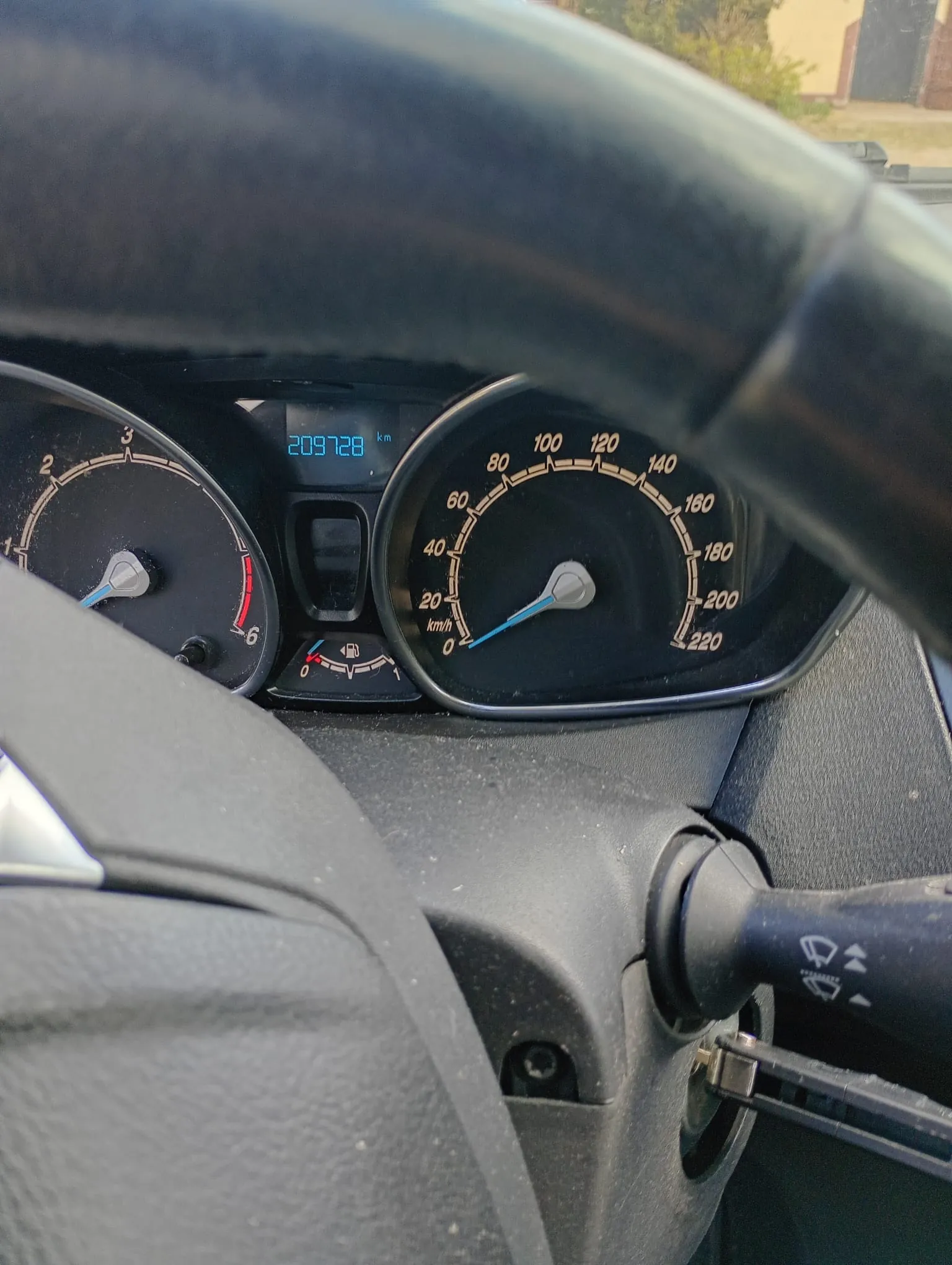Wnętrze samochodu z widoczną deską rozdzielczą. Po lewej stronie zegar obrotomieru z zakresem od 0 do 6, po prawej prędkościomierz z zakresem od 0 do 220 km/h. W centralnej części cyfrowy licznik przebiegu pokazujący 209728 km. Poniżej licznika przebiegu wskaźnik paliwa. Widoczna część kierownicy oraz dźwignia kierunkowskazów z symbolami. W tle przez przednią szybę fragment budynku i zieleni.