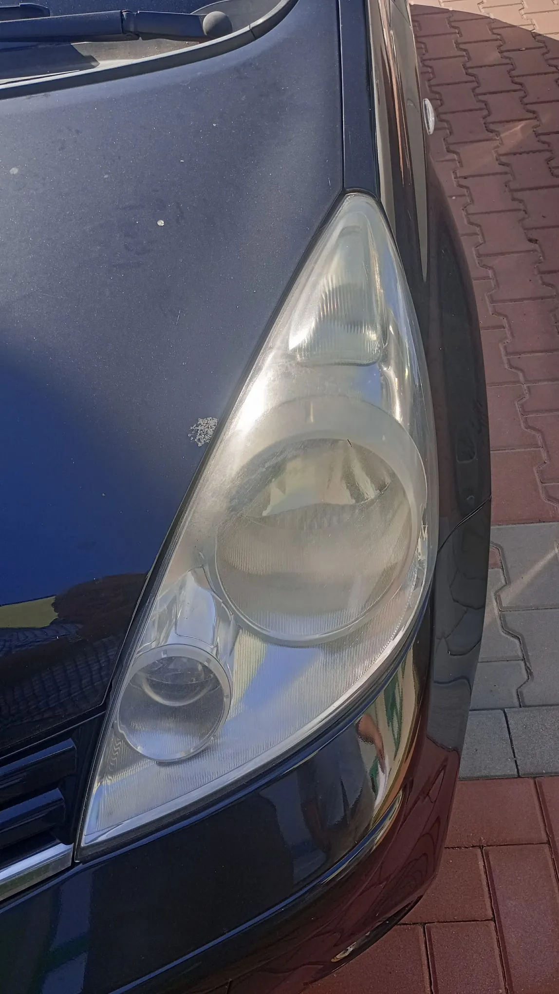 Zbliżenie na przedni prawy reflektor czarnego samochodu. Reflektor jest zmatowiały i utleniony. Obok lampy, na masce samochodu, widoczne jest niewielkie odpryśnięcie lakieru. Samochód zaparkowany jest na kostce brukowej w kolorze czerwonym i szarym.