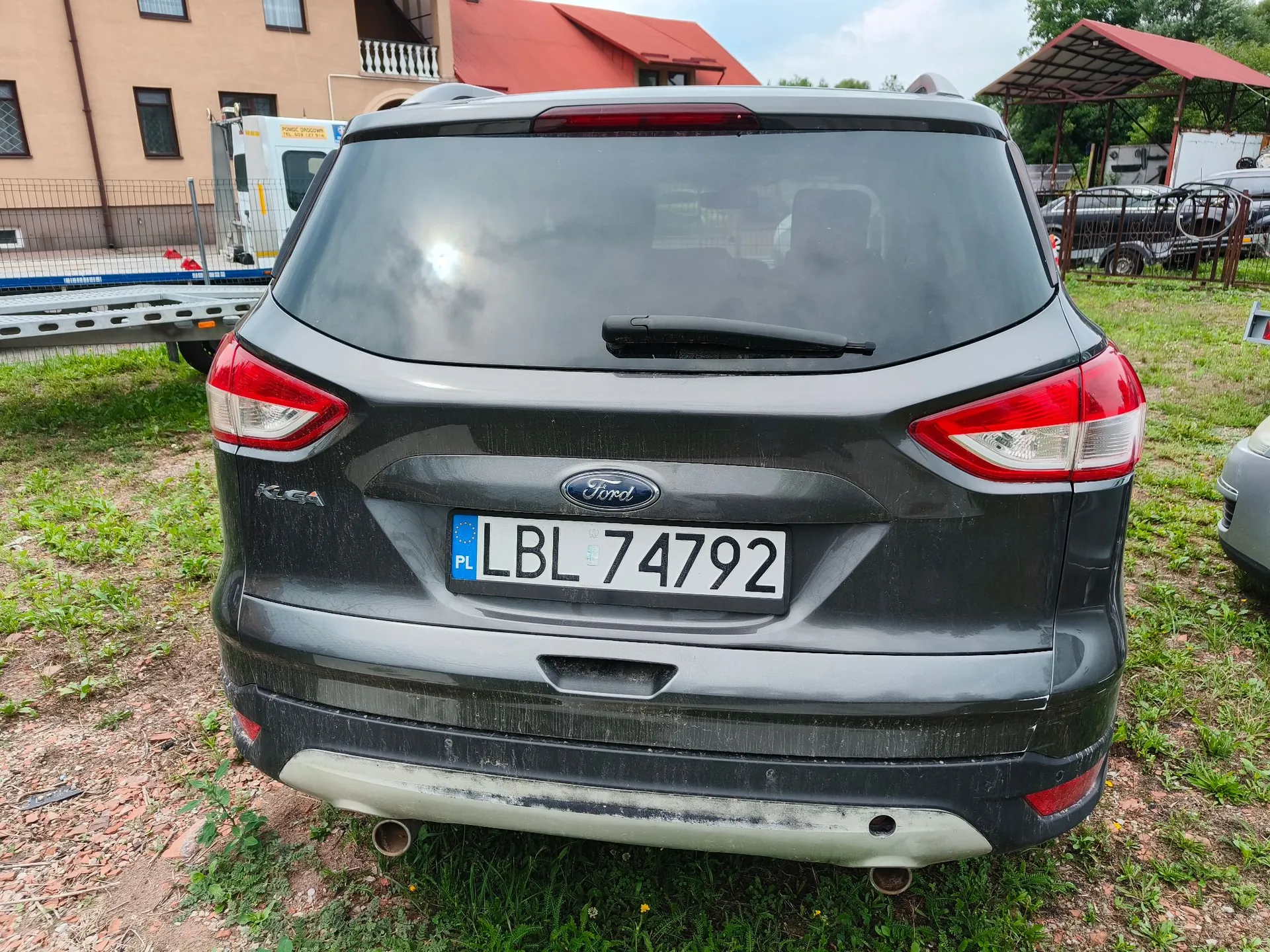 Tylny widok ciemnoszarego samochodu marki Ford. Pojazd posiada polską tablicę rejestracyjną o numerze LBL 74792. Samochód stoi na trawiastym terenie, w tle widoczne są budynki mieszkalne oraz metalowa konstrukcja z czerwonym dachem.