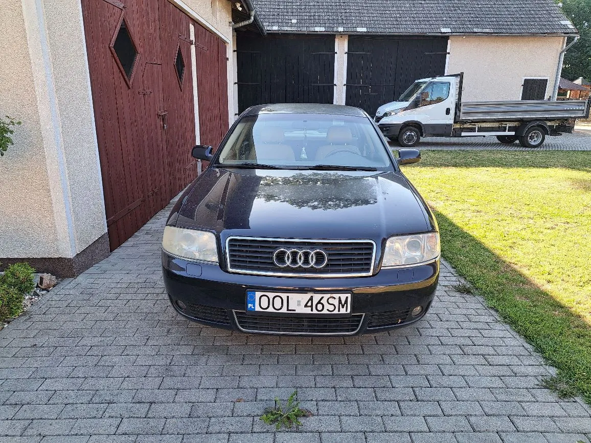Czarny samochód marki Audi zaparkowany przodem do widza na kostce brukowej. Samochód ma tablicę rejestracyjną o numerze OOL 46SM. W tle widoczne są brązowe drewniane drzwi garażowe po lewej stronie oraz biały samochód ciężarowy z otwartą paką zaparkowany na trawniku przed budynkiem z ciemnymi drzwiami.