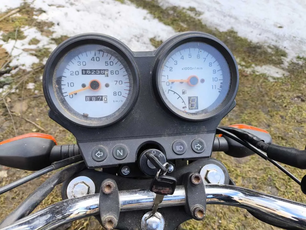 Zbliżenie na zegary motocykla. Po lewej stronie zegar prędkościomierza z zakresem 0-80 km/h, cyfrowym licznikiem przebiegu 16238 i licznikiem dziennym 887,0. Po prawej zegar obrotomierza z zakresem 0-11 x1000r/min, wskaźnikiem paliwa i cyfrowym wyświetlaczem wartości 8. Poniżej zegarów przyciski: strzałka w lewo, litera N, strzałka w prawo oraz dwa dodatkowe przyciski. Na kierownicy klucz zapłonu, lusterka boczne z pomarańczowymi elementami oraz metalowa rura kierownicy. Tło: trawa i śnieg.