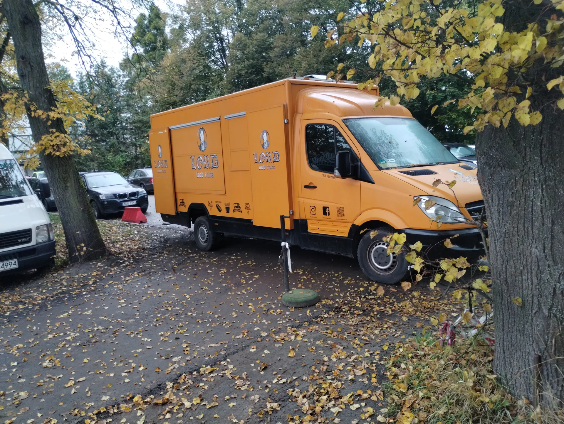 Pomarańczowy food truck z napisem "LORD food truck" na boku, zaparkowany na nawierzchni pokrytej żółtymi liśćmi. Po lewej stronie widoczny biały furgon z tablicą rejestracyjną 4994 oraz czarny samochód osobowy. Na boku food trucka znajdują się ikony jedzenia, logo mediów społecznościowych (Instagram, Facebook) oraz kod QR. W tle drzewa z żółtymi liśćmi, a na ziemi rozrzucone liście. Przy przednim kołem food trucka stoi zielony element przypominający podstawkę.