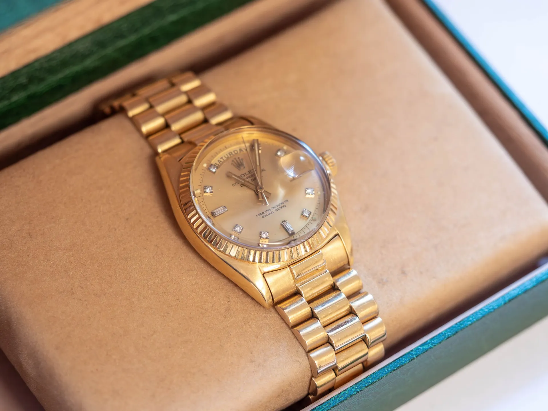 Złoty zegarek marki Rolex z bransoletą, leżący na beżowej poduszce wewnątrz zielonego pudełka. Tarcza zegarka jest jasna z widocznym tekstem: 'SATURDAY', 'ROLEX OYSTER PERPETUAL', 'SUPERLATIVE CHRONOMETER OFFICIALLY CERTIFIED'. Zegarek posiada złoty bezel w kształcie ząbków oraz diamentowe indeksy godzinowe.