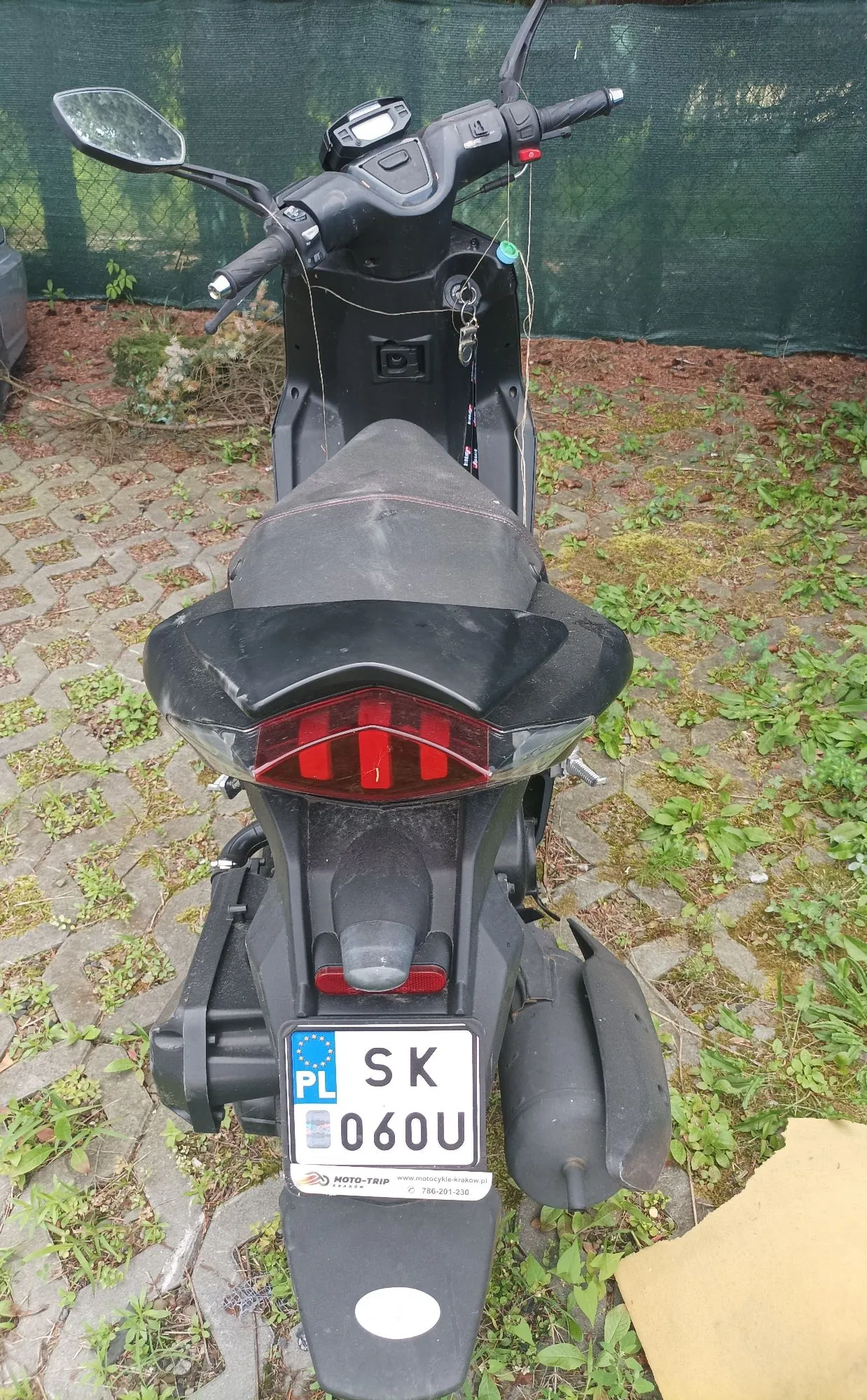 Czarny skuter zaparkowany tyłem do widza na kostce brukowej z przerostami trawy. Widoczny jest tylny reflektor, siedzenie oraz tablica rejestracyjna o numerze SK 060U z oznaczeniem PL. Poniżej tablicy znajduje się naklejka z napisem 'MOTO-TRIP' i numerem telefonu '786-201-238'. W tle widać zieloną siatkę ogrodzeniową.