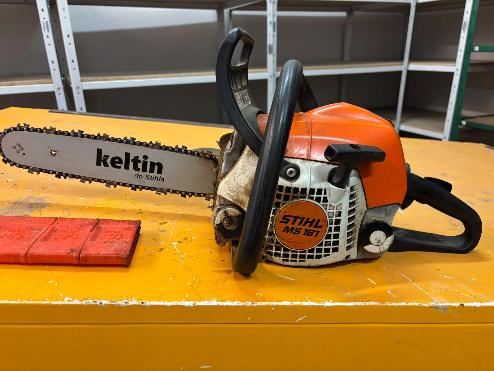 Pomarańczowo-biała piła spalinowa marki Stihl, model MS 181, leżąca na żółtym blacie. Na prowadnicy piły znajduje się napis 'keltin do Stihla'. Obok piły leży czerwony, prostokątny przedmiot. W tle widoczne są białe regały magazynowe.