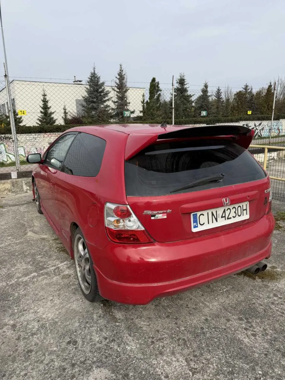 Czerwony samochód Honda hatchback z widocznym tyłem, tablicą rejestracyjną CIN 4230H z flagą PL, napisem Sport i logo Hondy. Samochód stoi na betonowej powierzchni, w tle siatka ogrodzeniowa z żółtymi znakami numerowanymi 74, 75, 76, 77, budynek, drzewa iglaste oraz pomalowane graffiti ściany. Niebo jest pochmurne, a na samochodzie widoczny jest czarny spoiler i podwójne wydechy.