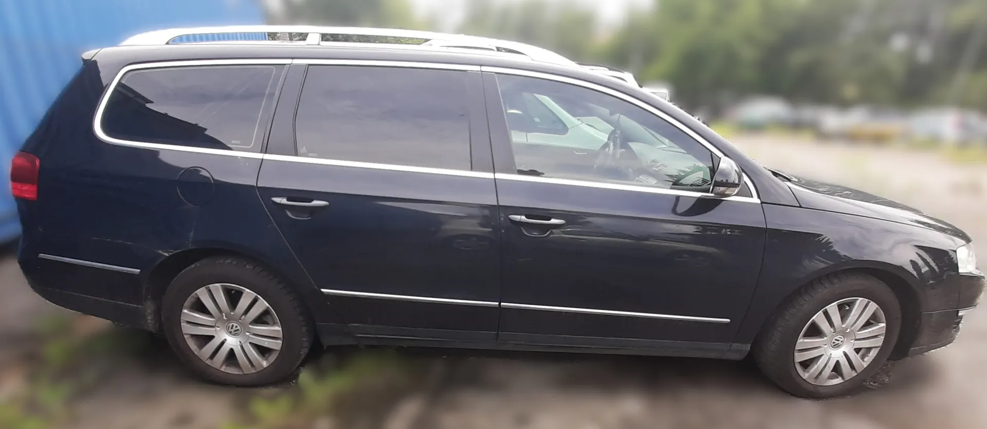 Ciemnoniebieski samochód osobowy typu kombi marki Volkswagen z białym dachowym bagażnikiem. Pojazd posiada przyciemniane szyby, srebrne felgi z logo VW oraz chromowane listwy nad i pod oknami. W tle rozmyte drzewa i niebieska konstrukcja. Samochód zaparkowany na nierównym terenie z widoczną trawą.
