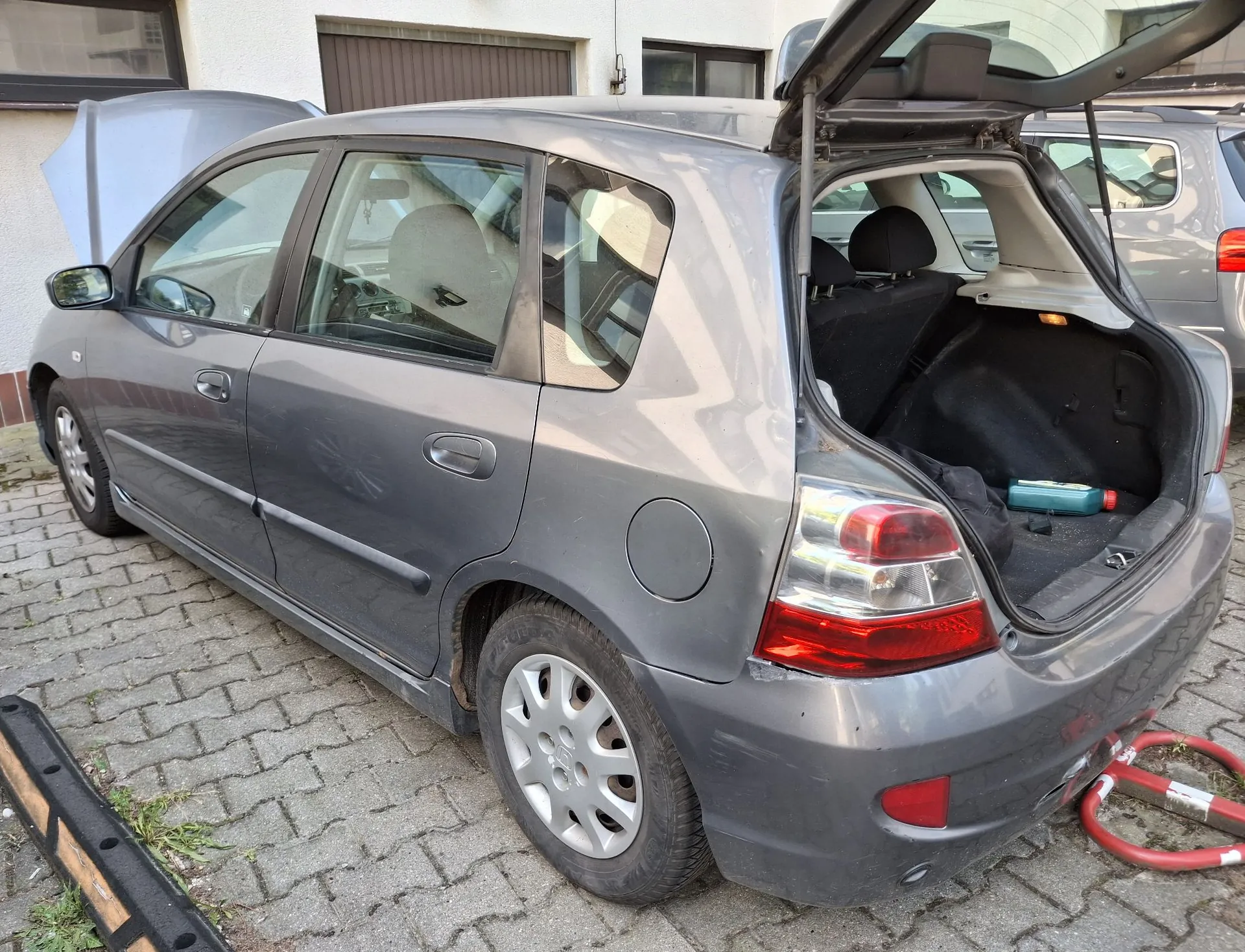 Szary samochód osobowy typu hatchback zaparkowany na kostce brukowej. Pojazd ma otwartą maskę silnika oraz otwartą klapę bagażnika, w którym znajduje się niebieska butelka z płynem. Samochód jest widoczny z profilu od strony pasażera, w tle znajduje się biały budynek z oknami i bramą garażową.