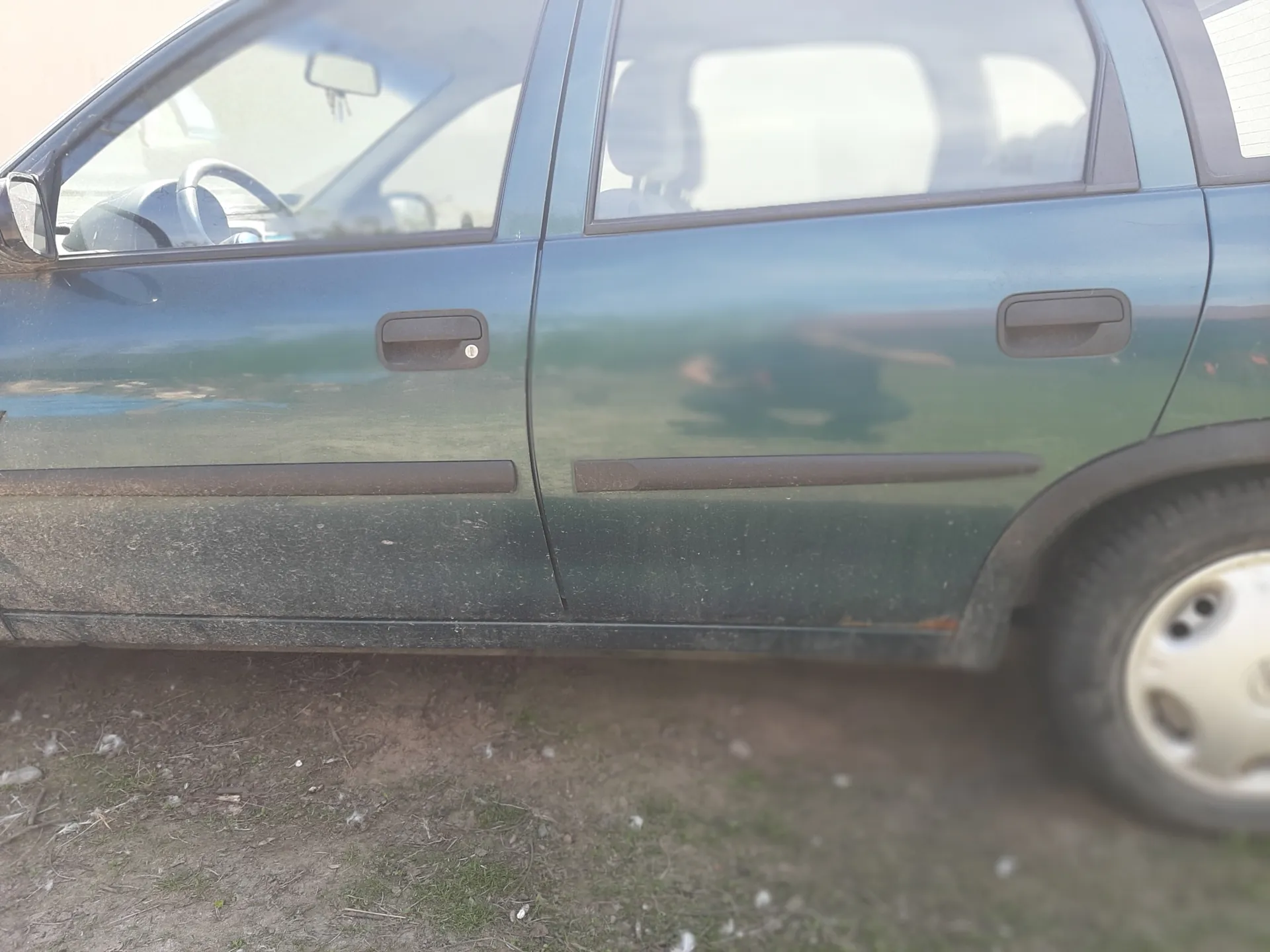 Boczny widok niebieskiego samochodu osobowego z dwoma widocznymi drzwiami. Samochód ma brudną karoserię, szczególnie w dolnej części. Widoczne są uchwyty drzwiowe, lusterko boczne oraz fragment wnętrza przez przednią szybę, w tym kierownica. Pod samochodem znajduje się ziemia z rzadką trawą i drobnymi śmieciem. Brak czytelnego tekstu na obrazie.