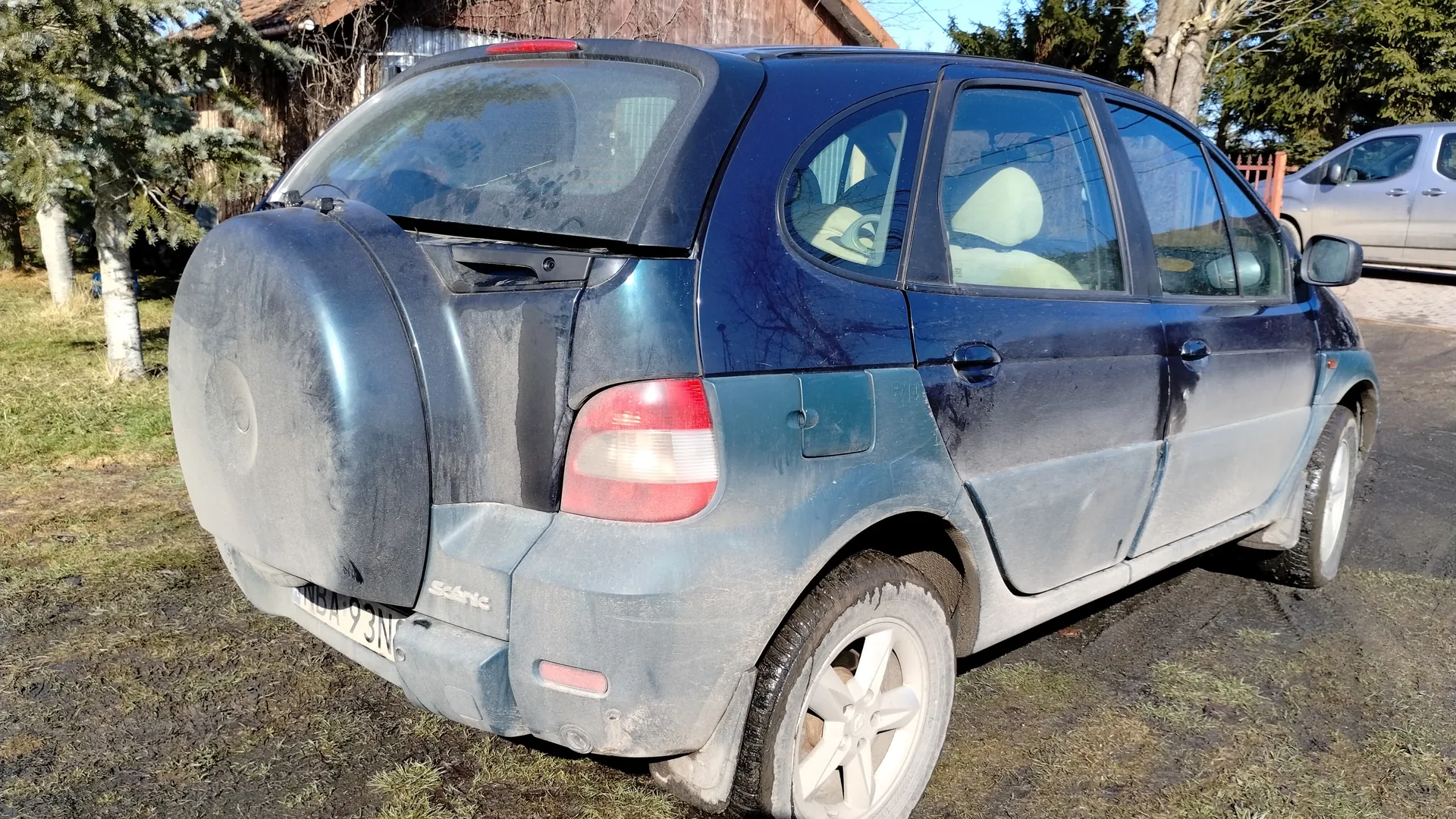 Niebieski samochód osobowy z widocznym z tyłu kołem zapasowym, brudnym nadwoziem i tablicą rejestracyjną NBA 93N. W tle drewniany budynek, drzewa oraz srebrny samochód zaparkowany na podjeździe. Samochód stoi na trawie i błotnistym terenie.