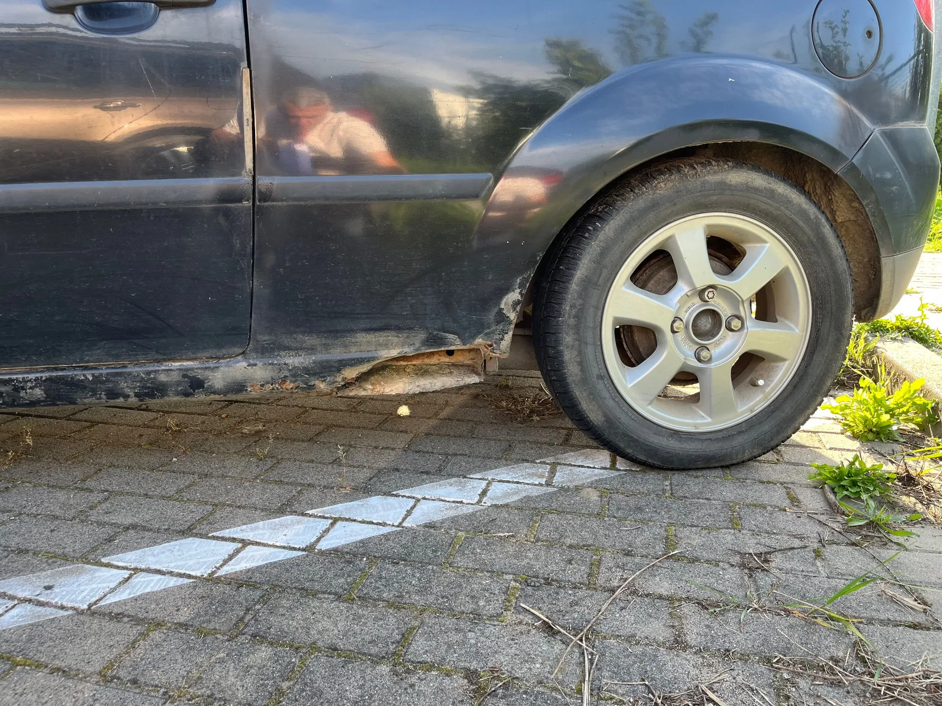 Zbliżenie na dolną część czarnego samochodu, pokazujące tylne koło z aluminiową felgą oraz znaczne uszkodzenia i korozję progu nadwozia. Samochód stoi na kostce brukowej z namalowaną białą linią.