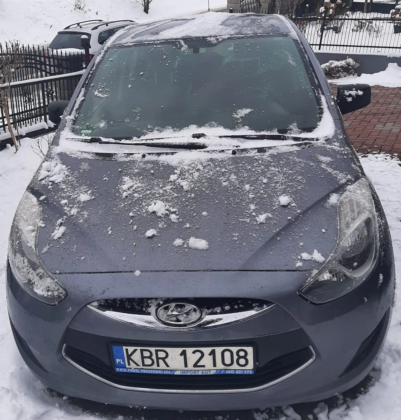 Szary samochód Hyundai z widocznym emblematem, pokryty śniegiem na masce, przedniej szybie, dachu i reflektorach. Tablica rejestracyjna KBR 12108 z oznaczeniem PL. W tle czarny metalowy płot, biały samochód oraz śnieżna sceneria z czerwonym brukiem.