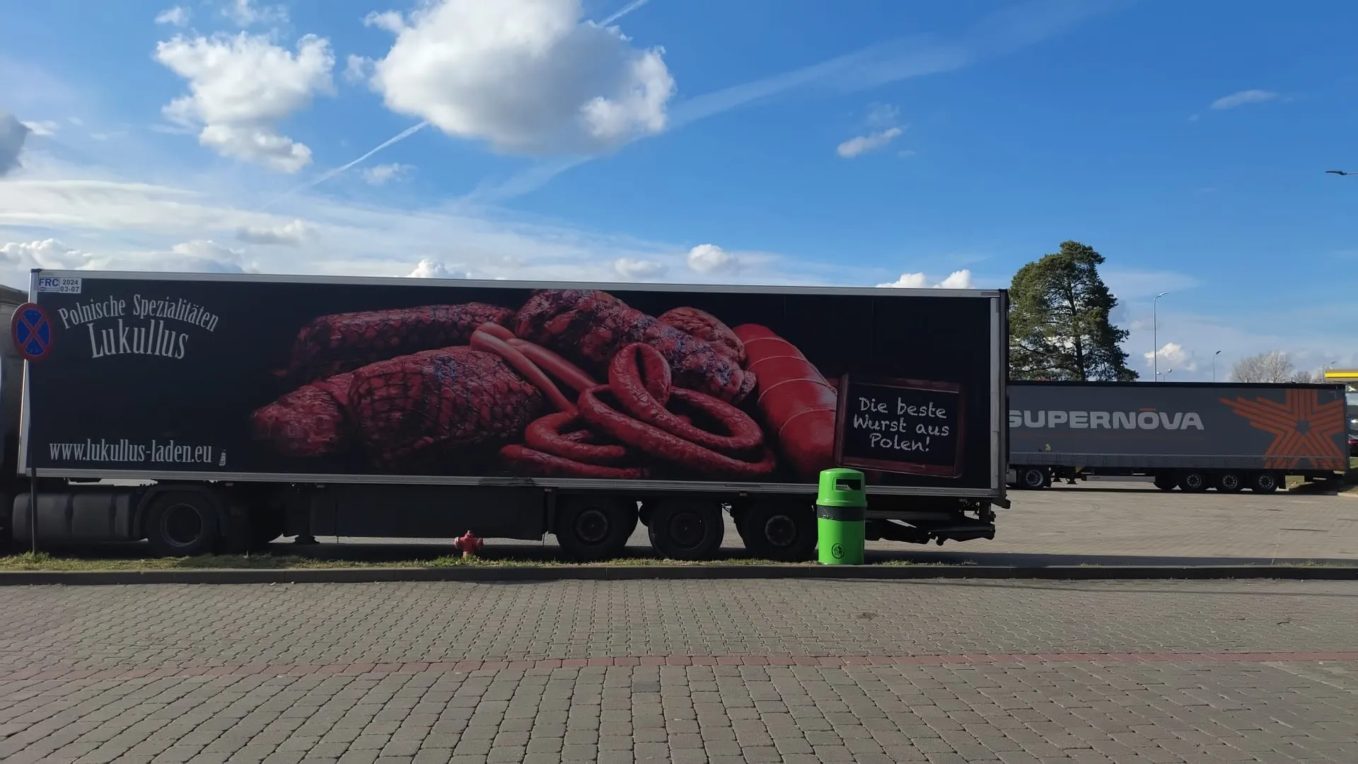 Duży naczepa ciężarówki z reklamą przedstawiającą różne rodzaje kiełbas i mięsnych specjałów na czarnym tle. Na lewo widoczny napis 'Polnische Spezialitäten Lukullus' oraz adres strony internetowej 'www.lukullus-laden.eu'. Po prawej stronie tabliczka z napisem 'Die beste Wurst aus Polen!'. W tle druga naczepa z napisem 'SUPERNOVA', zielony kosz na śmieci, czerwony hydrant, znak drogowy oraz niebo z chmurami. Podłoga wyłożona ceglanymi płytami.