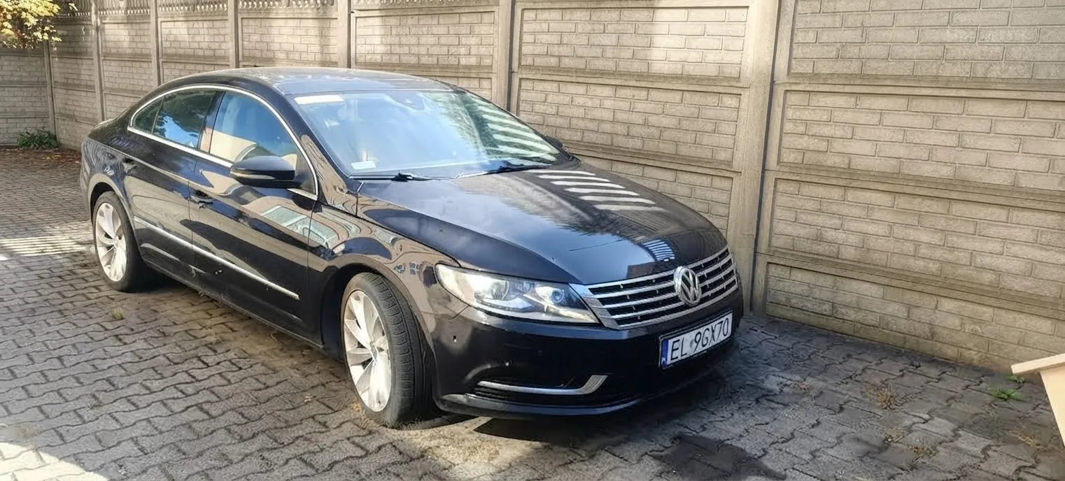 Czarny samochód osobowy marki Volkswagen zaparkowany na kostce brukowej obok jasnego, ceglanego muru. Pojazd jest ustawiony pod kątem, widoczny z przodu i z boku. Tablica rejestracyjna samochodu ma numer EL 96GX70.