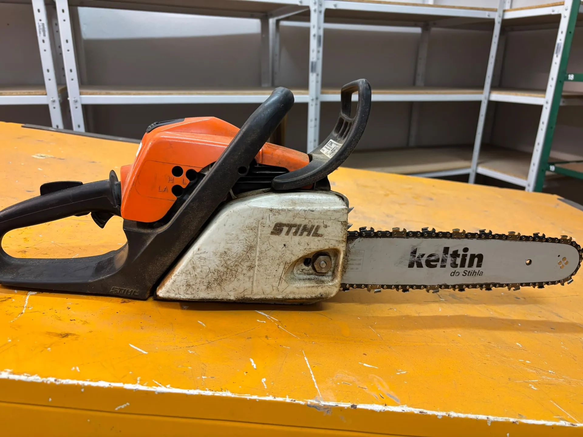 Pomarańczowo-biała piła łańcuchowa marki Stihl leżąca na żółtym blacie. Na prowadnicy łańcucha widnieje napis 'keltin do Stihla'. W tle znajdują się metalowe regały magazynowe.