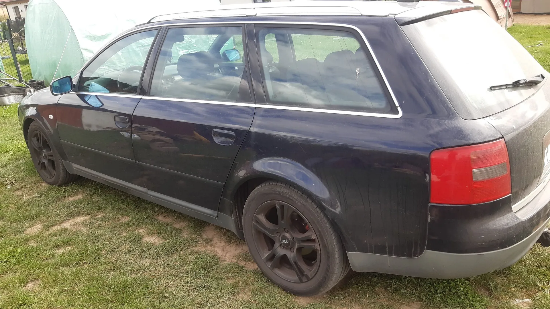 Granatowy samochód typu kombi zaparkowany na trawie. Widoczny jest bok i tył pojazdu z ciemnymi felgami aluminiowymi. W tle znajduje się jasny namiot ogrodowy.