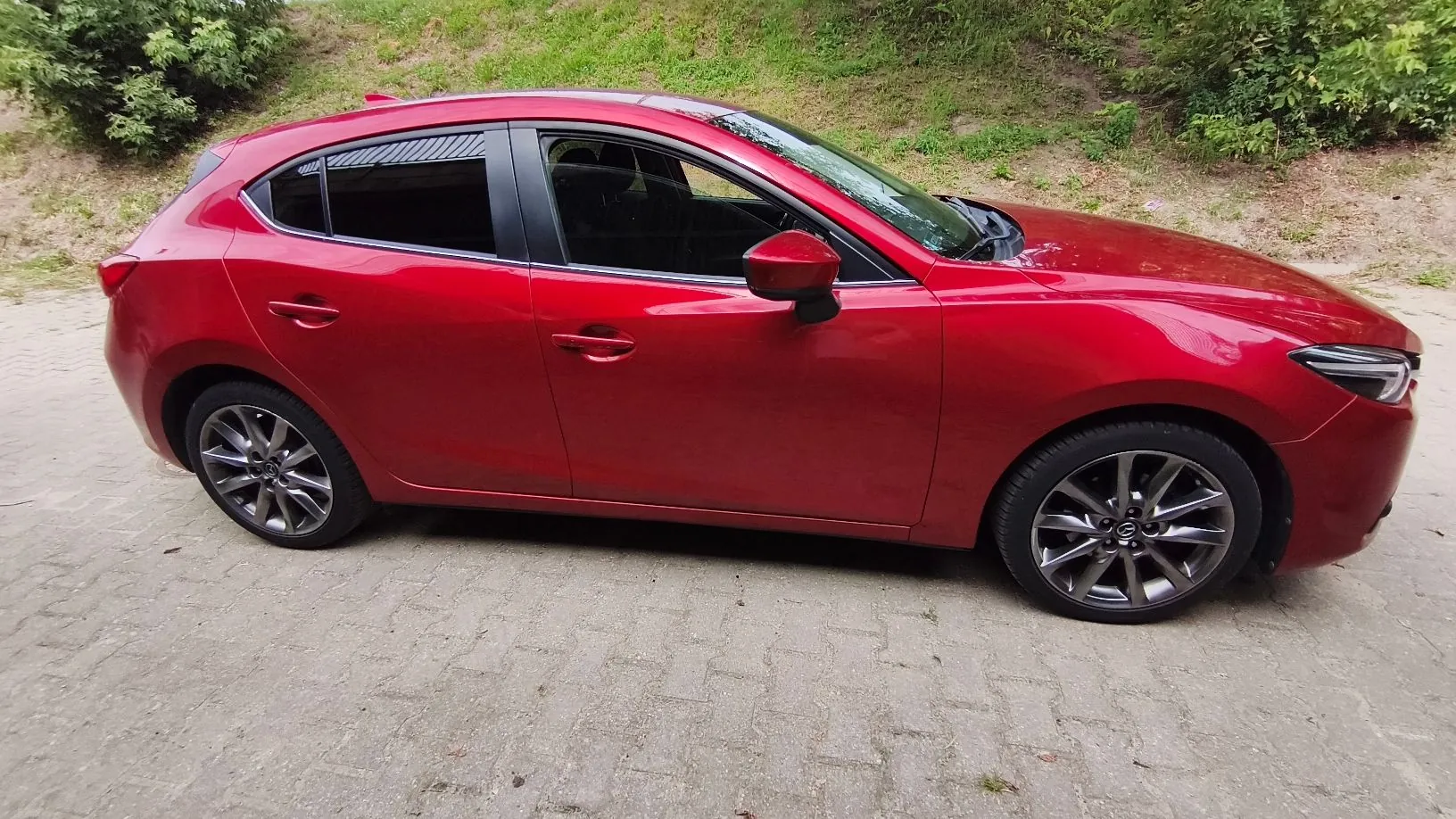 Profil czerwonego samochodu osobowego typu hatchback zaparkowanego na kostce brukowej na tle zielonej roślinności.