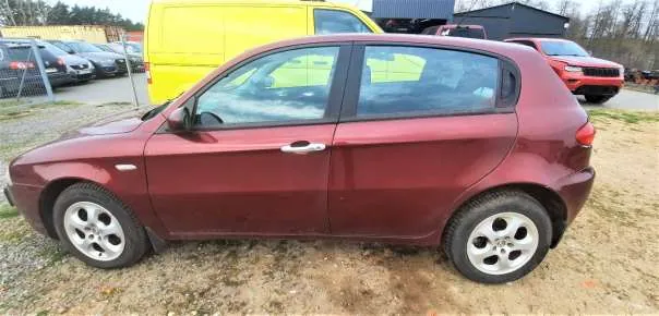 Czerwony samochód osobowy typu hatchback zaparkowany na żwirowym terenie. W tle widoczne są inne pojazdy: żółty van, czerwony SUV oraz samochody za ogrodzeniem. W oddali drzewa i budynki. Samochód ma srebrne felgi i czarne opony.