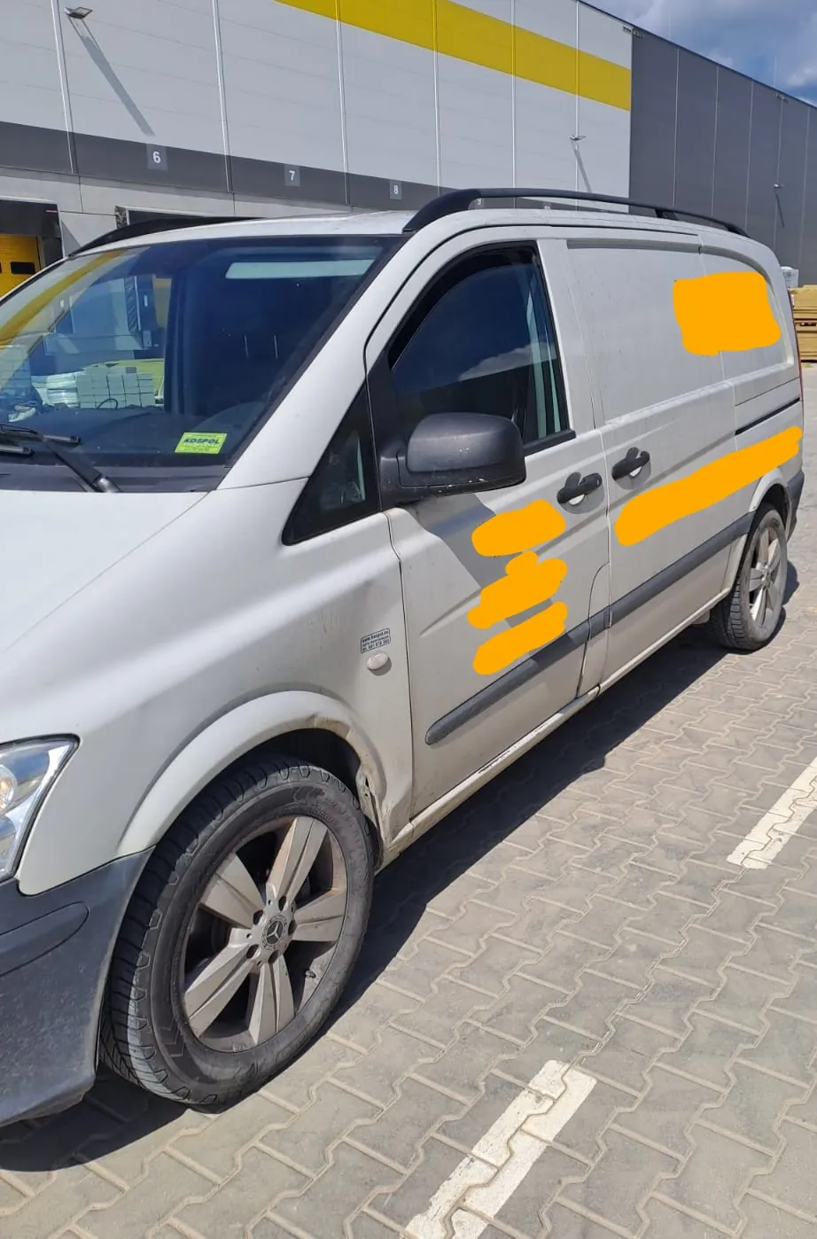 Srebrny samochód dostawczy zaparkowany na kostce brukowej przed budynkiem magazynowym. Pojazd jest widoczny z boku, z przodu i z profilu. Część nadwozia, w tym drzwi pasażera i panel boczny, jest zakryta pomarańczowymi plamami maskującymi.