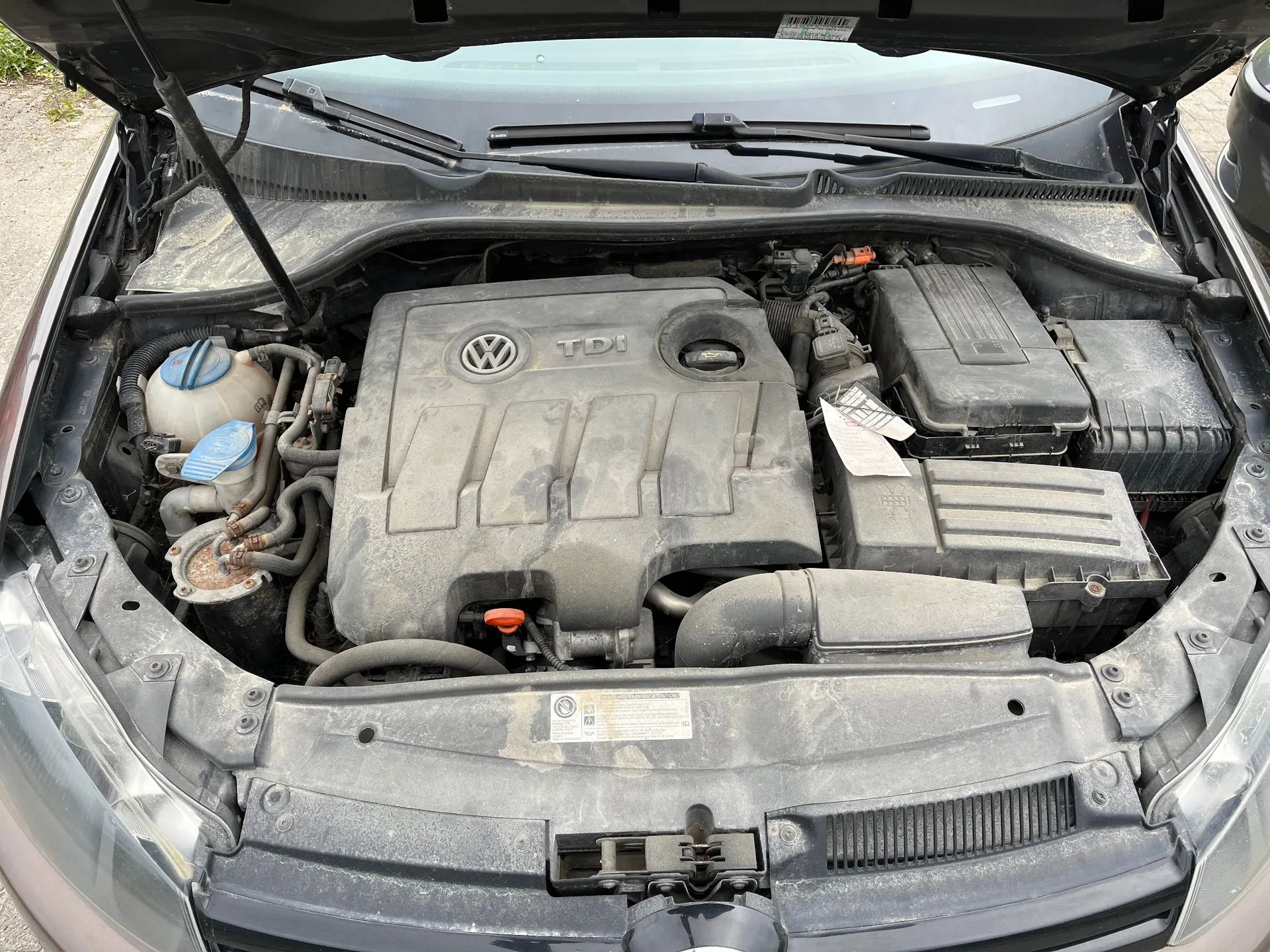Widok z góry na otwartą maskę samochodu, ukazujący zakurzony silnik z plastikową osłoną z logo Volkswagena i napisem TDI. Po lewej stronie widoczny jest zbiornik z płynem spryskiwaczy z niebieską zakrętką, a po prawej skrzynka bezpieczników i obudowa filtra powietrza.