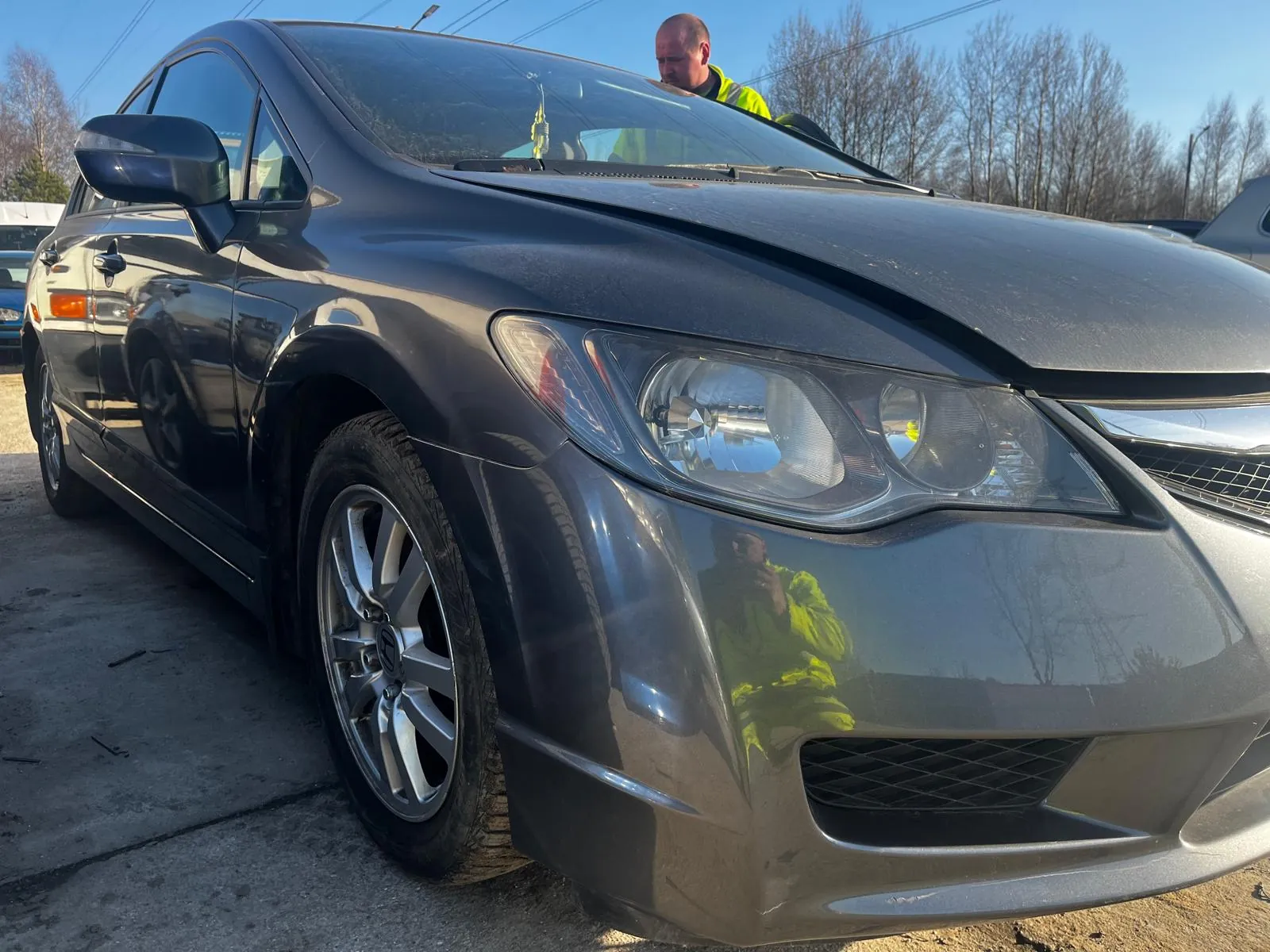 Ciemnoszary sedan zaparkowany na betonowej powierzchni. Osoba w jasnożółtej kurtce odblaskowej stoi za samochodem, blisko przedniej szyby. Tło z bezlistnymi drzewami i jasnym niebem. Na betonie widoczne są pęknięcia i drobne szczątki.