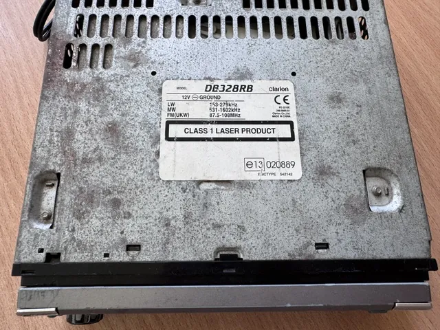 Zdjęcie przedstawiające tylny panel srebrnego urządzenia elektronicznego marki Clarion, prawdopodobnie odtwarzacza samochodowego. Na obudowie znajduje się biała naklejka z tekstem: 'MODEL DB328RB clarion', '12V GROUND', 'LW 153-279kHz', 'MW 531-1602kHz', 'FM(UKW) 87.5-108MHz', 'CLASS 1 LASER PRODUCT' oraz oznaczenia 'CE', 'e13 020889' i 'FACTORY 542142'. Górna część obudowy posiada otwory wentylacyjne.