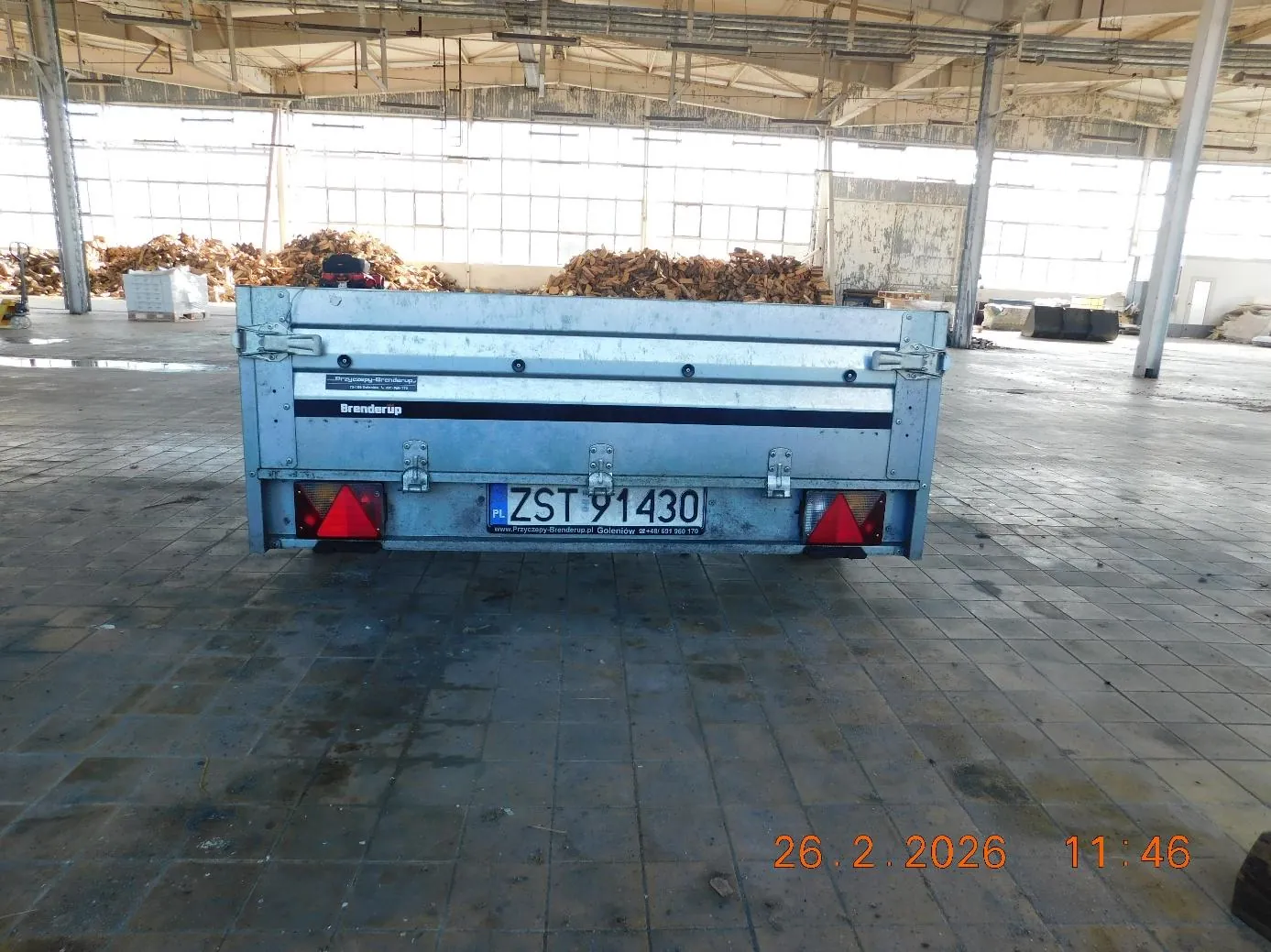 Tył metalowego przyczepu lub pojazdu z tablicą rejestracyjną ZST 91430, napisem Brenderup oraz czerwonymi trójkątnymi światłami. Pojazd znajduje się w przestronnym hali przemysłowej z betonową podłogą, wysokim sufitem i dużymi oknami. W tle widoczne są stosy drewna i elementy infrastruktury hali. W prawym dolnym rogu obrazu znajduje się pomarańczowy znaczek z datą 26.2.2026 i godziną 11:46.
