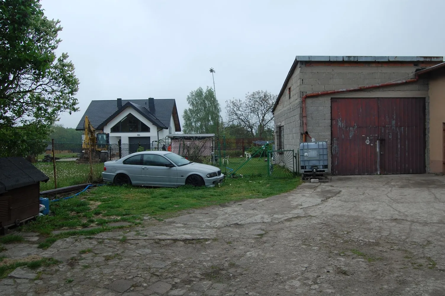 Zdjęcie przedstawia podwórze z betonowym placem na pierwszym planie. Po lewej stronie stoi srebrny samochód osobowy zaparkowany na trawie. W tle widoczny jest nowoczesny biały dom z ciemnym dachem oraz szary budynek gospodarczy z dużymi, czerwonymi, metalowymi drzwiami garażowymi. Obok garażu stoi plastikowy zbiornik na wodę. Po lewej stronie znajduje się żółta koparka oraz zielone drzewa.