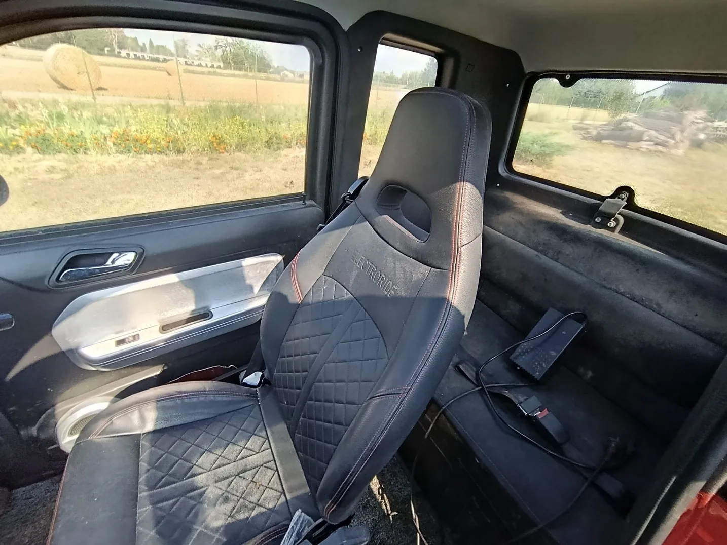 Wnętrze pojazdu z widokiem na czarny, sportowy fotel z przeszyciami w romby i czerwonymi detalami. Na oparciu fotela widnieje napis 'ELECTRORIDE'. W tle widać okno z widokiem na pole z belami siana oraz tylną część kabiny z czarnym elementem przypominającym zasilacz i kablem.